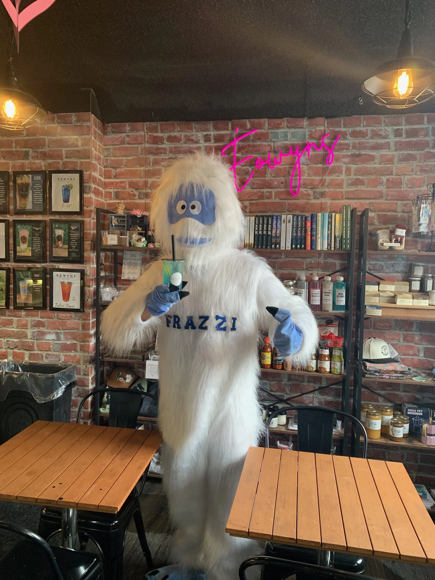 A person dressed in a large white furry costume resembling a yeti or Abominable Snowman, with a blue face and big eyes, stands inside a café or shop. The costume has the word 'FRAZZI' embroidered on the chest. The mannequin is holding a colorful drink in one hand and pointing with the other. Behind, there are brick walls, books, and shelves with various products. A pink neon sign reads 'Fowyns'.