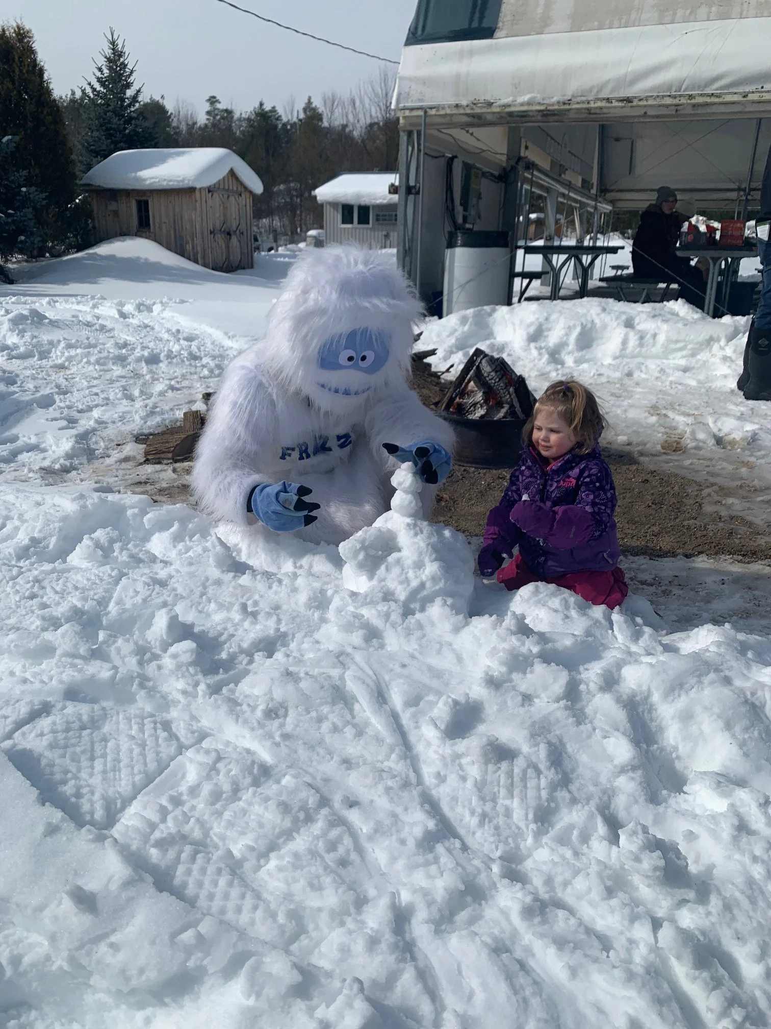 A person dressed in a furry white Yetti costume playing with a young girl in purple winter clothing as they build a snowman outdoors in a snowy area. In the background, there is a tent, a small shed, and a person sitting at a table.