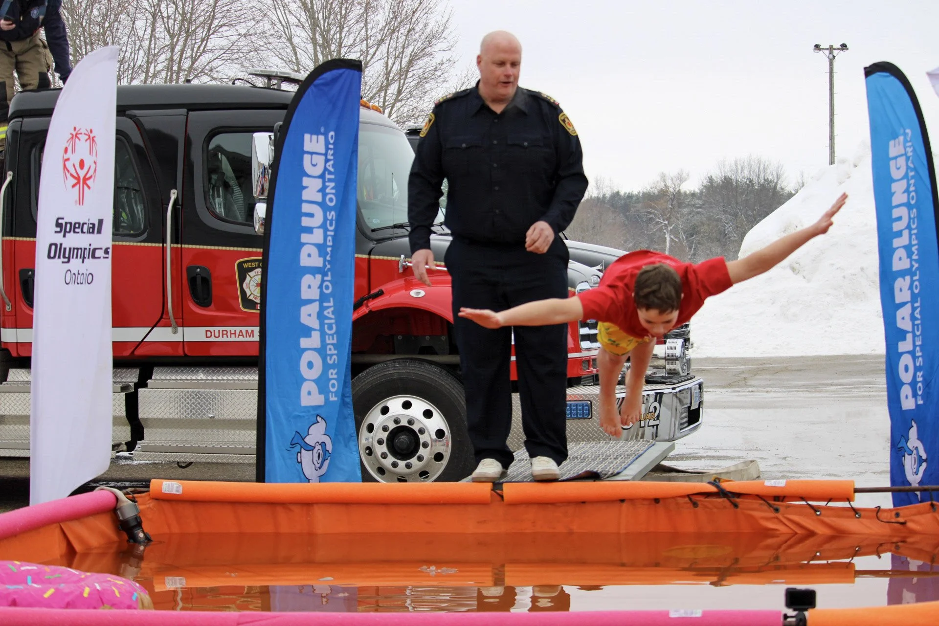 A child in yellow shorts and red shirt diving into a body of water at a Special Olympics event, overseen by a police officer, with flags and emergency vehicles in the background.