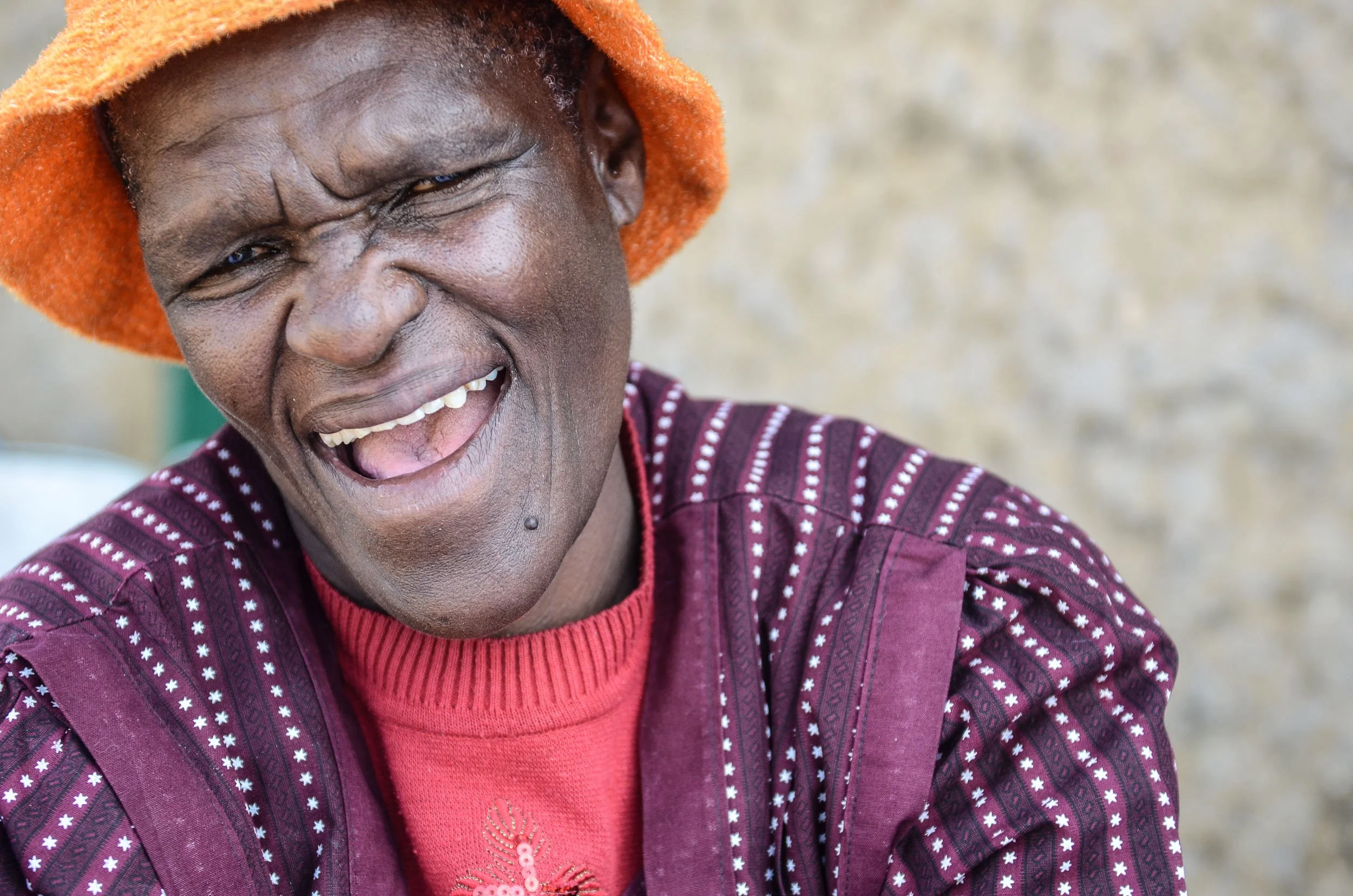 A smiling man wearing an orange hat and a maroon and white patterned shirt, with a red knitted sweater underneath, standing outdoors in front of a beige wall.