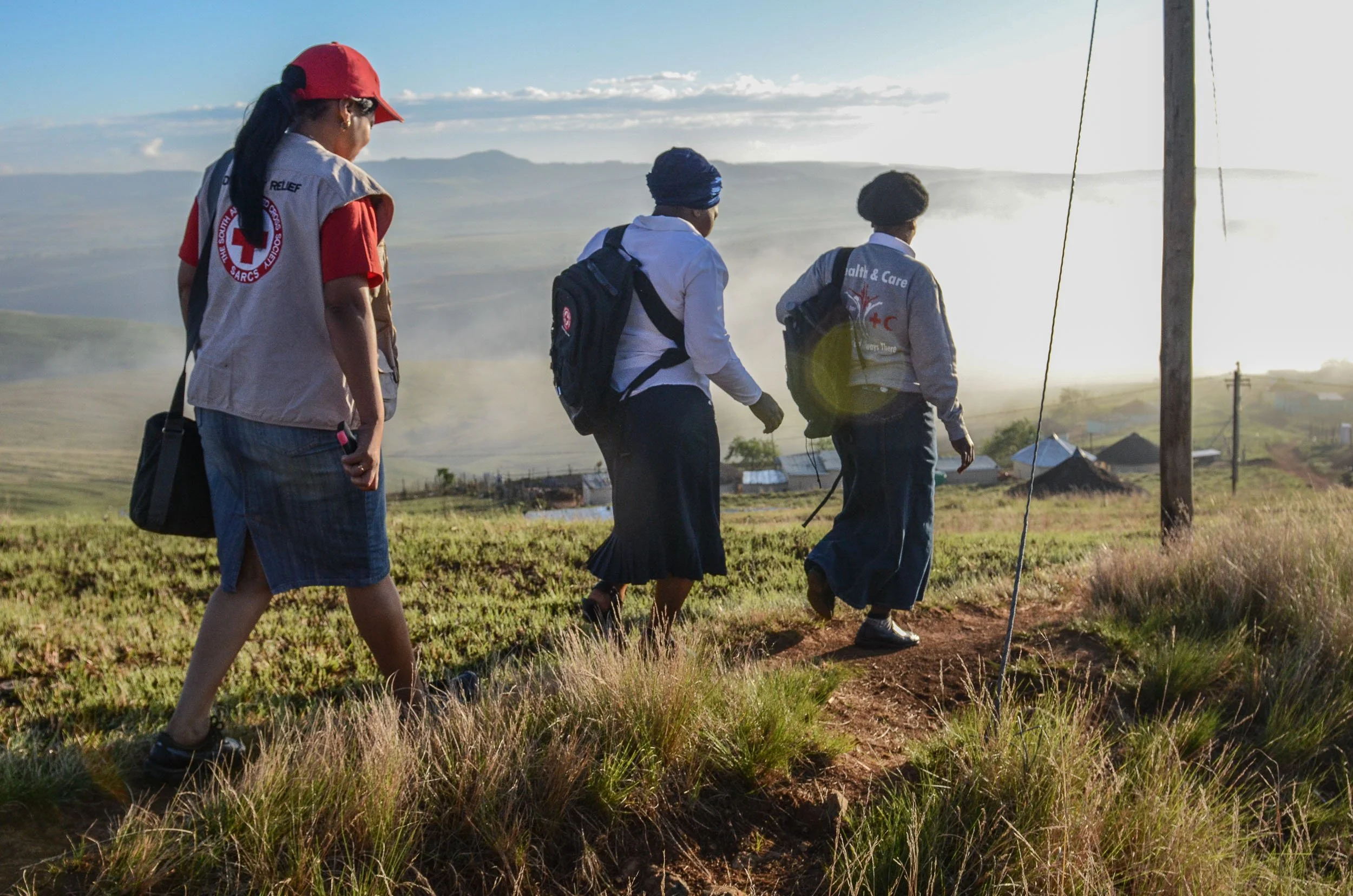Group of Red Cross aid workers walking through grassy field towards a small village in a rural area with hills and mountains in the background, during sunrise or sunset.