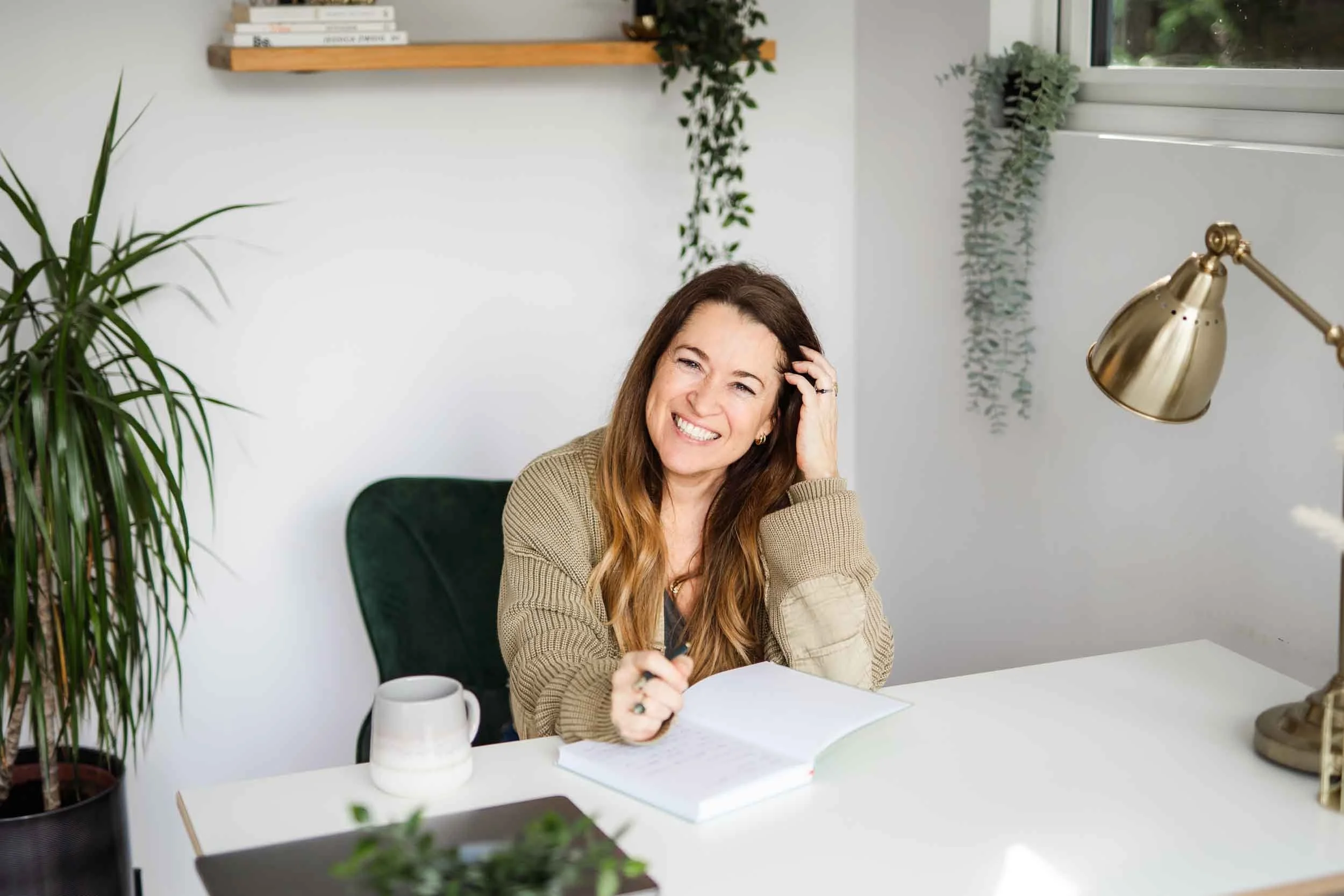 A woman sitting at a white desk, smiling and holding a pen, with an open notebook in front of her. There is a large green plant on the left and a gold desk lamp on the right. The background includes a white wall with a high shelf holding books and some hanging plants near a window.