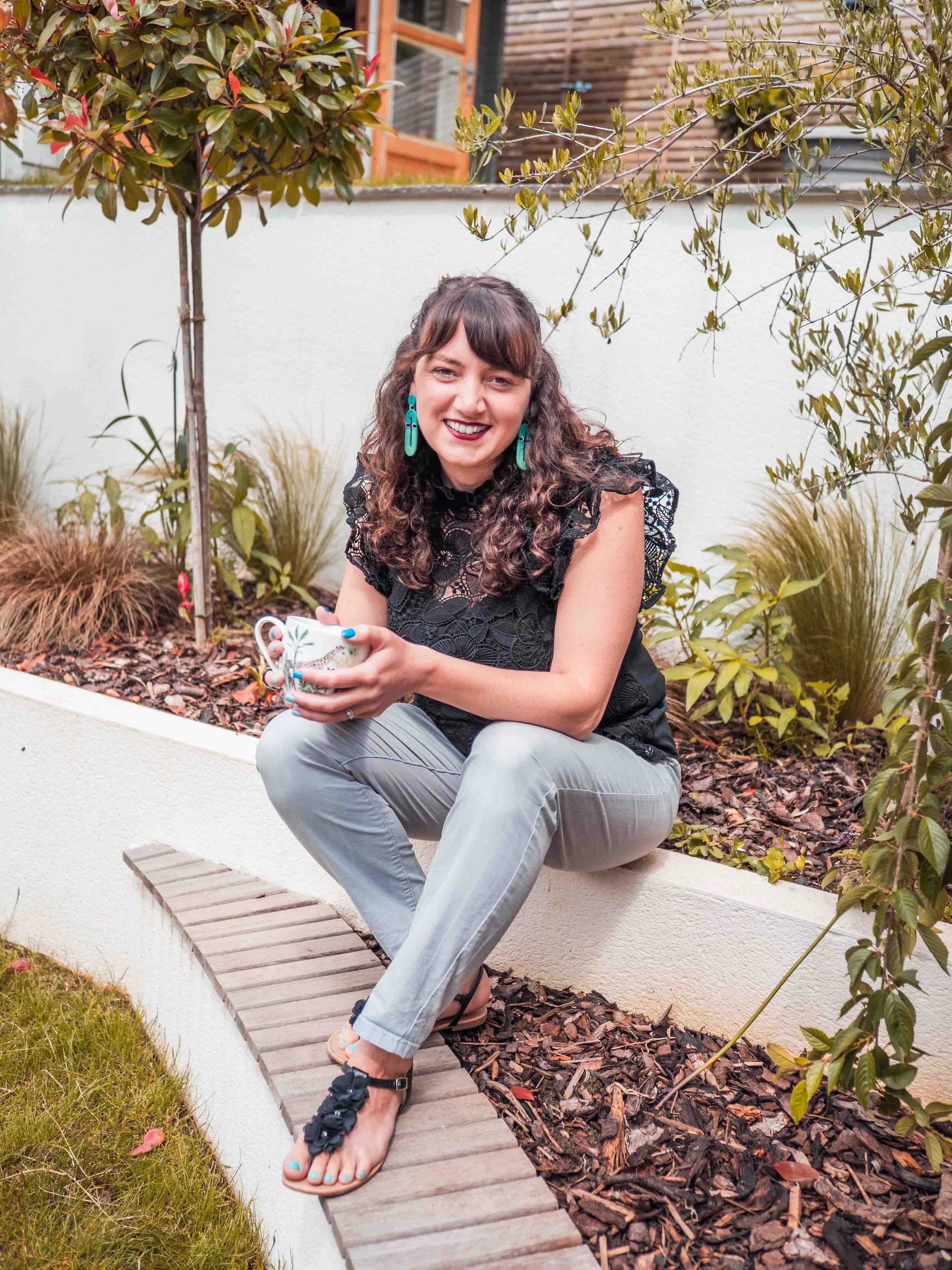 A woman with curly brown hair, wearing a black lace top, gray pants, and black sandals, sitting outdoors on a garden wall, holding a mug, and smiling.