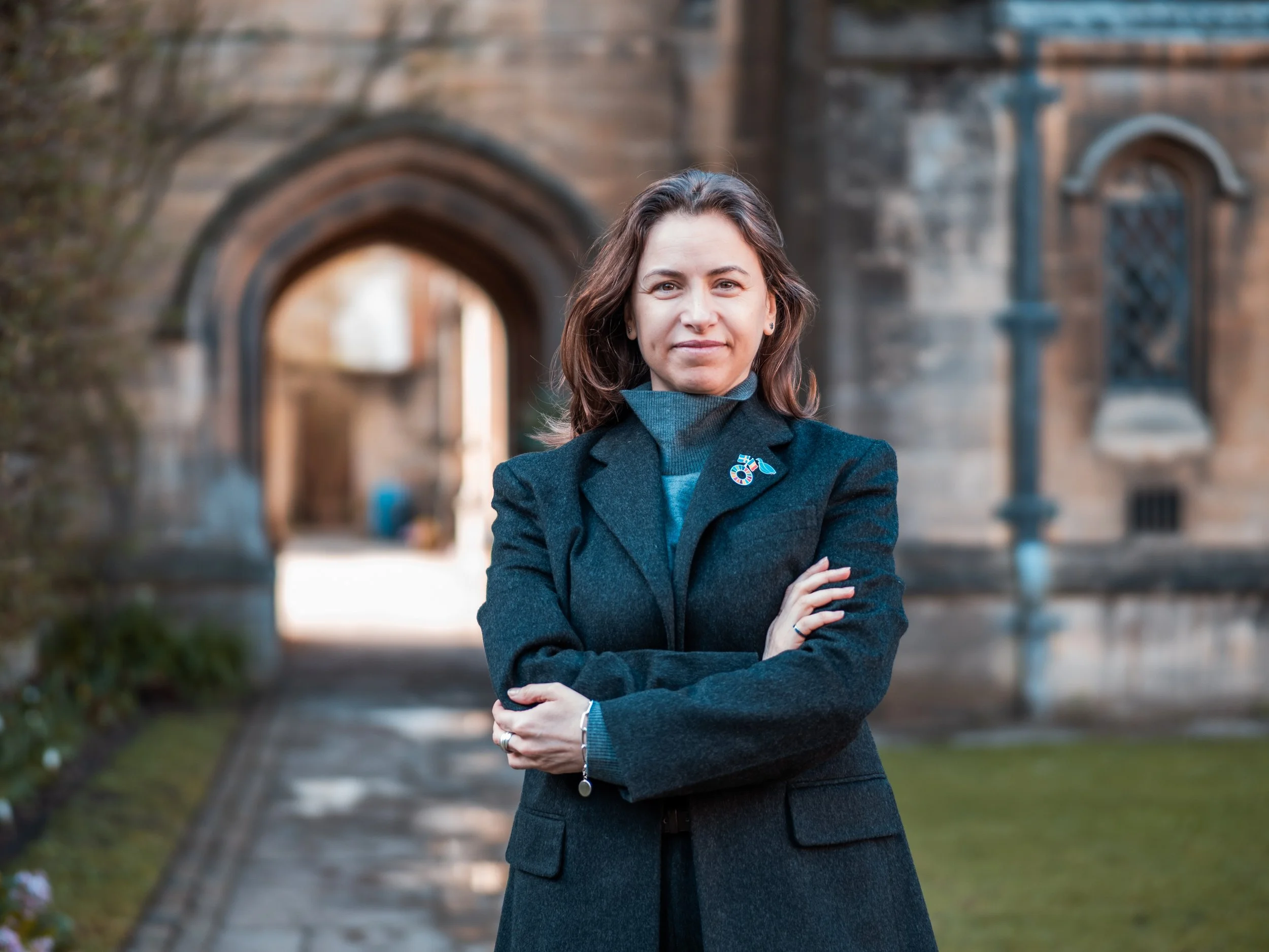A woman standing outdoors in front of an arched stone gateway, wearing a dark blazer with a lapel pin, with her arms crossed.