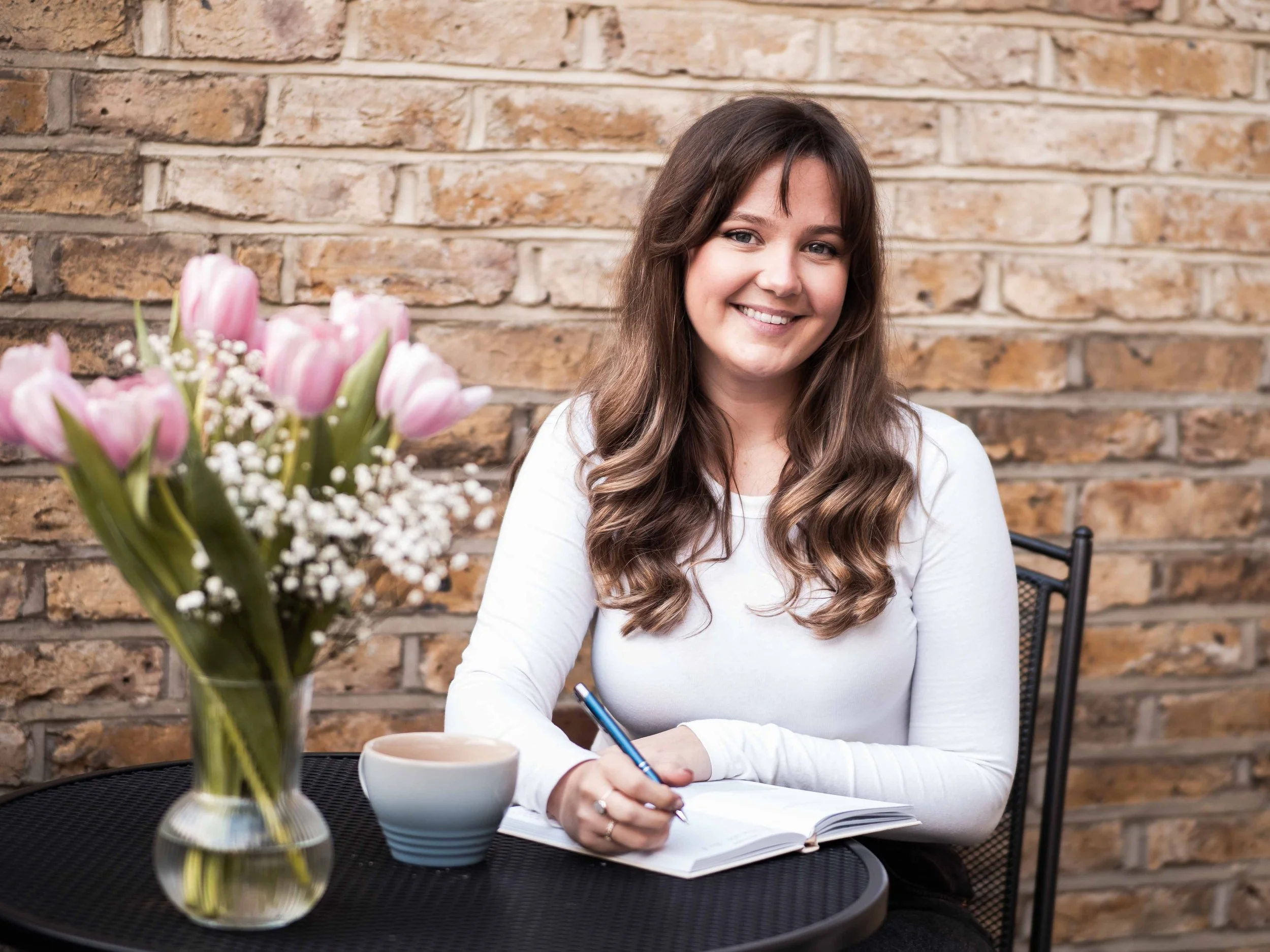 A woman with long brown hair smiling, sitting at a table with a notebook, pen, mug, and a vase of pink tulips with white baby's breath flowers, against a brick wall background.