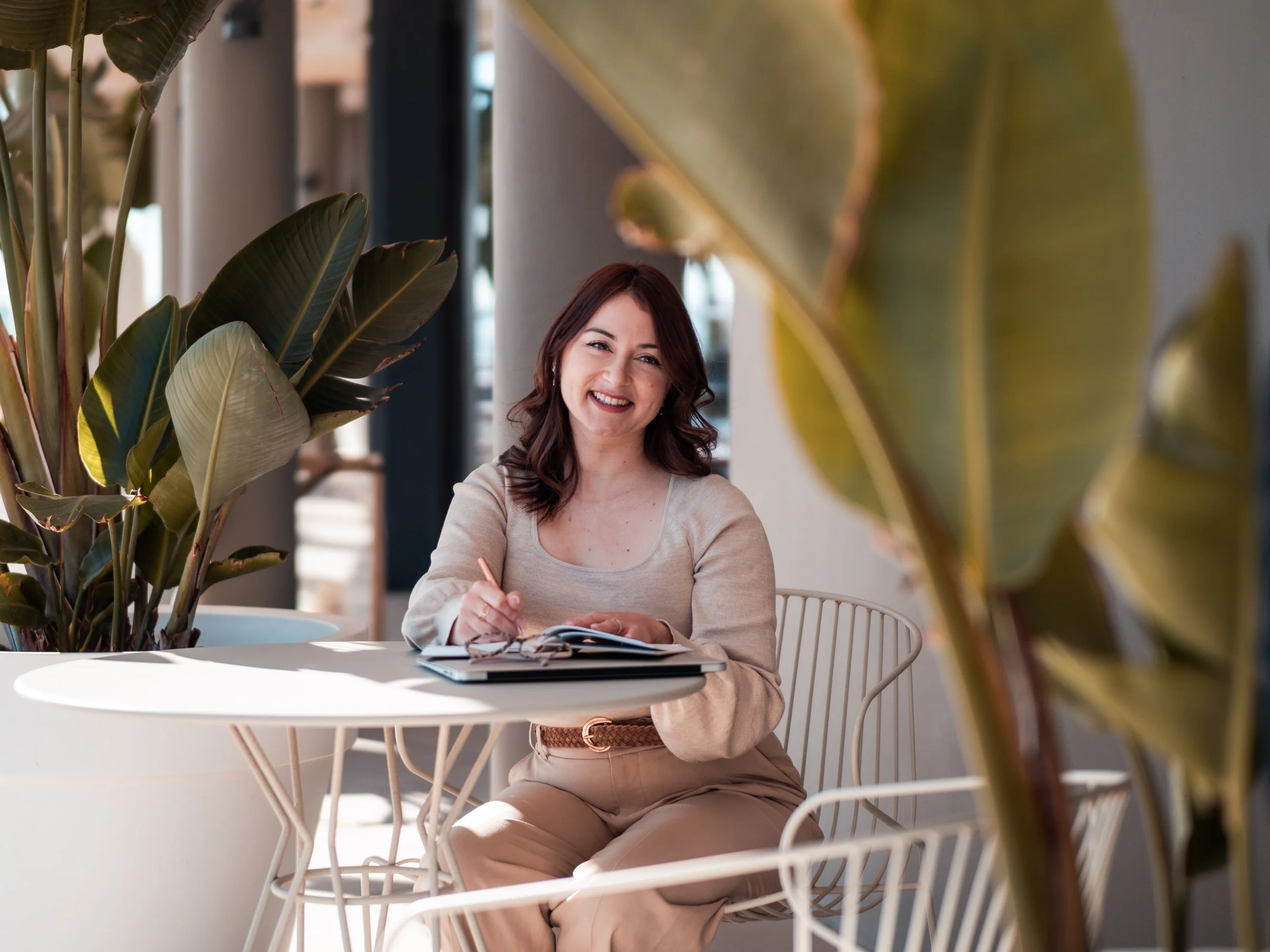 A woman with brown hair smiling while sitting at a white table, writing in a notebook, surrounded by large green plants in a bright, modern indoor space.