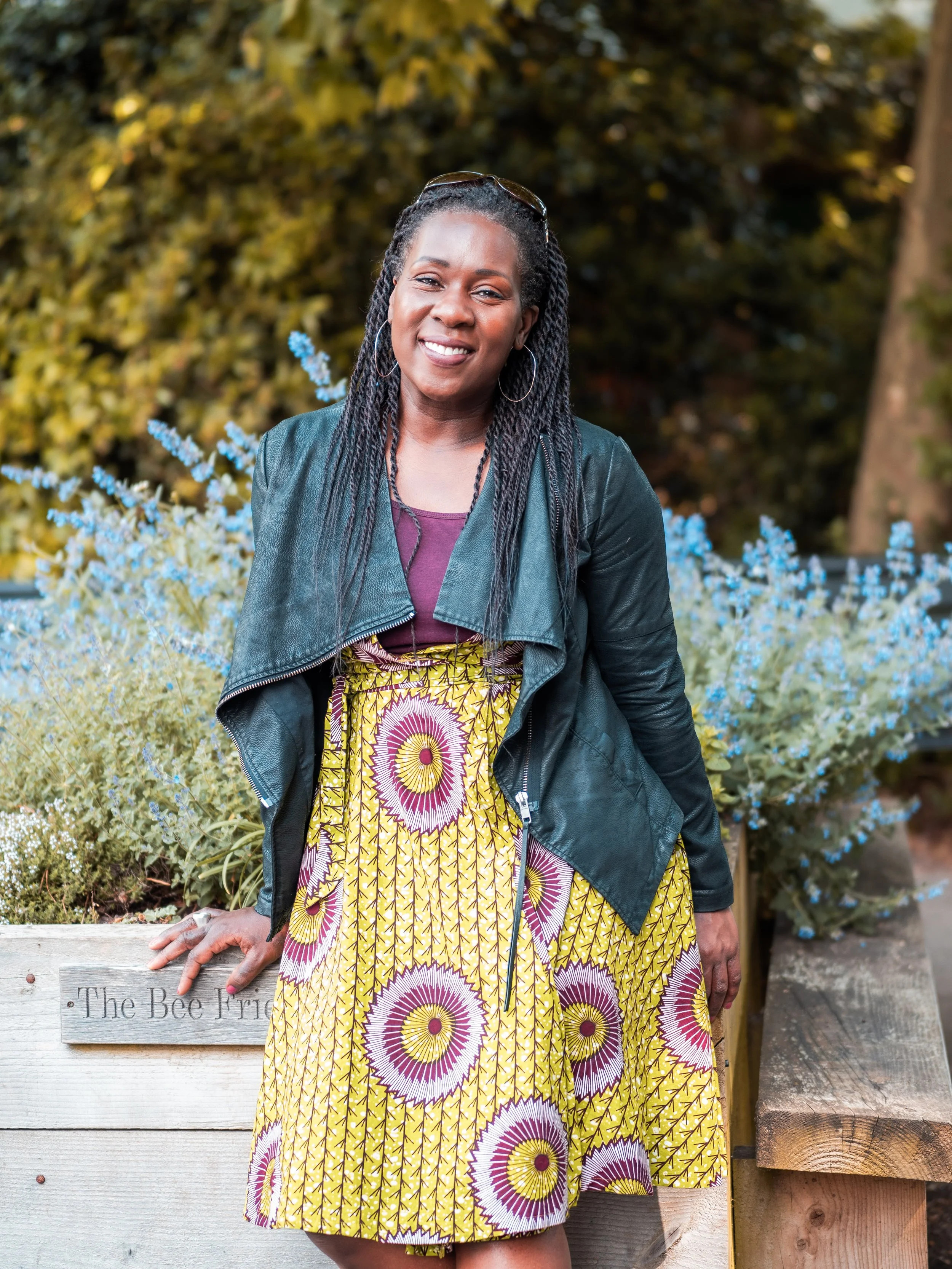 A woman with braided hair and hoop earrings posing outdoors in front of blue flowers and green foliage, wearing a leather jacket and a colorful patterned skirt, smiling at the camera.