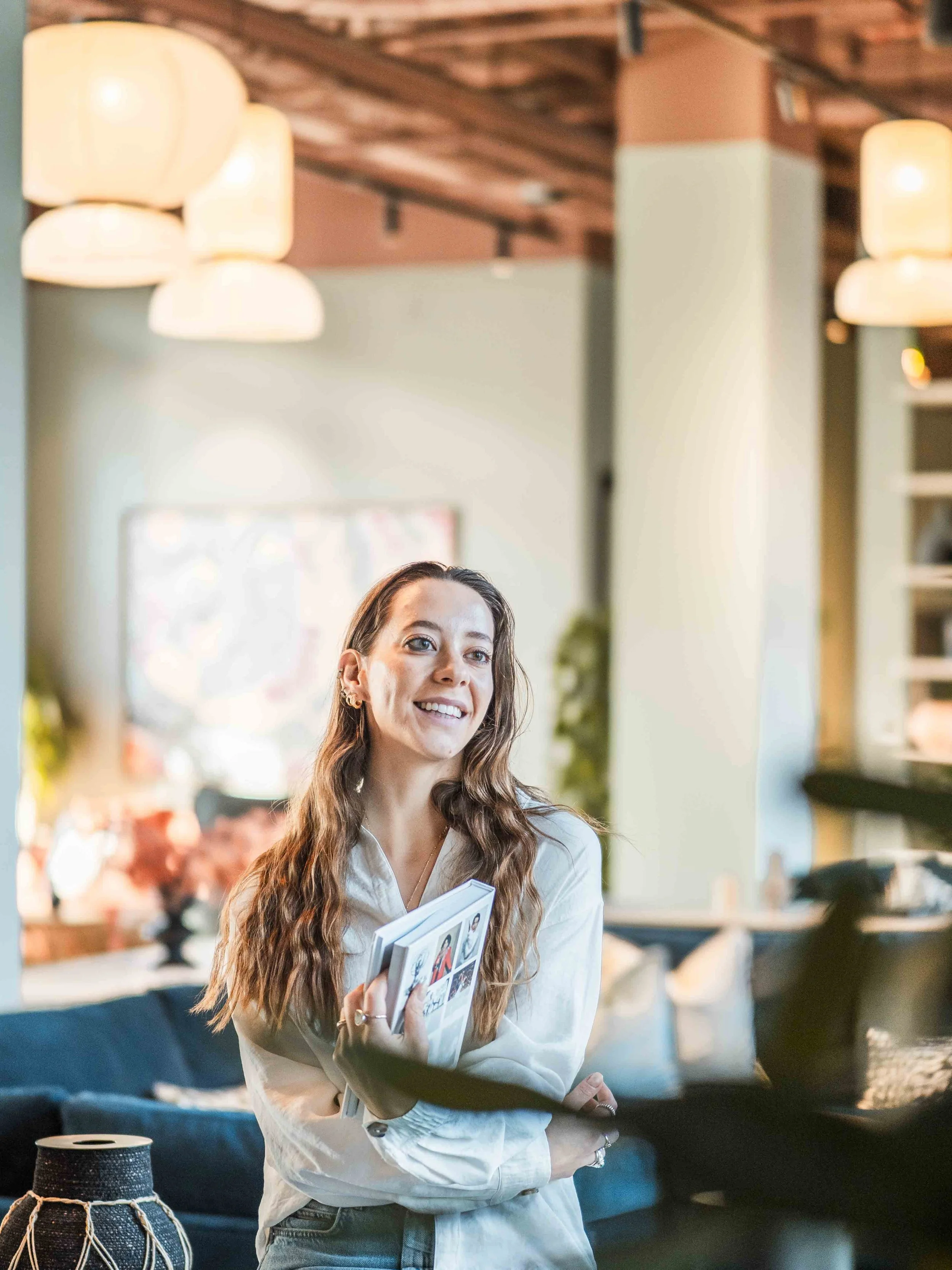 A woman with long wavy hair holding a magazine or brochure in a well-lit, modern interior space.