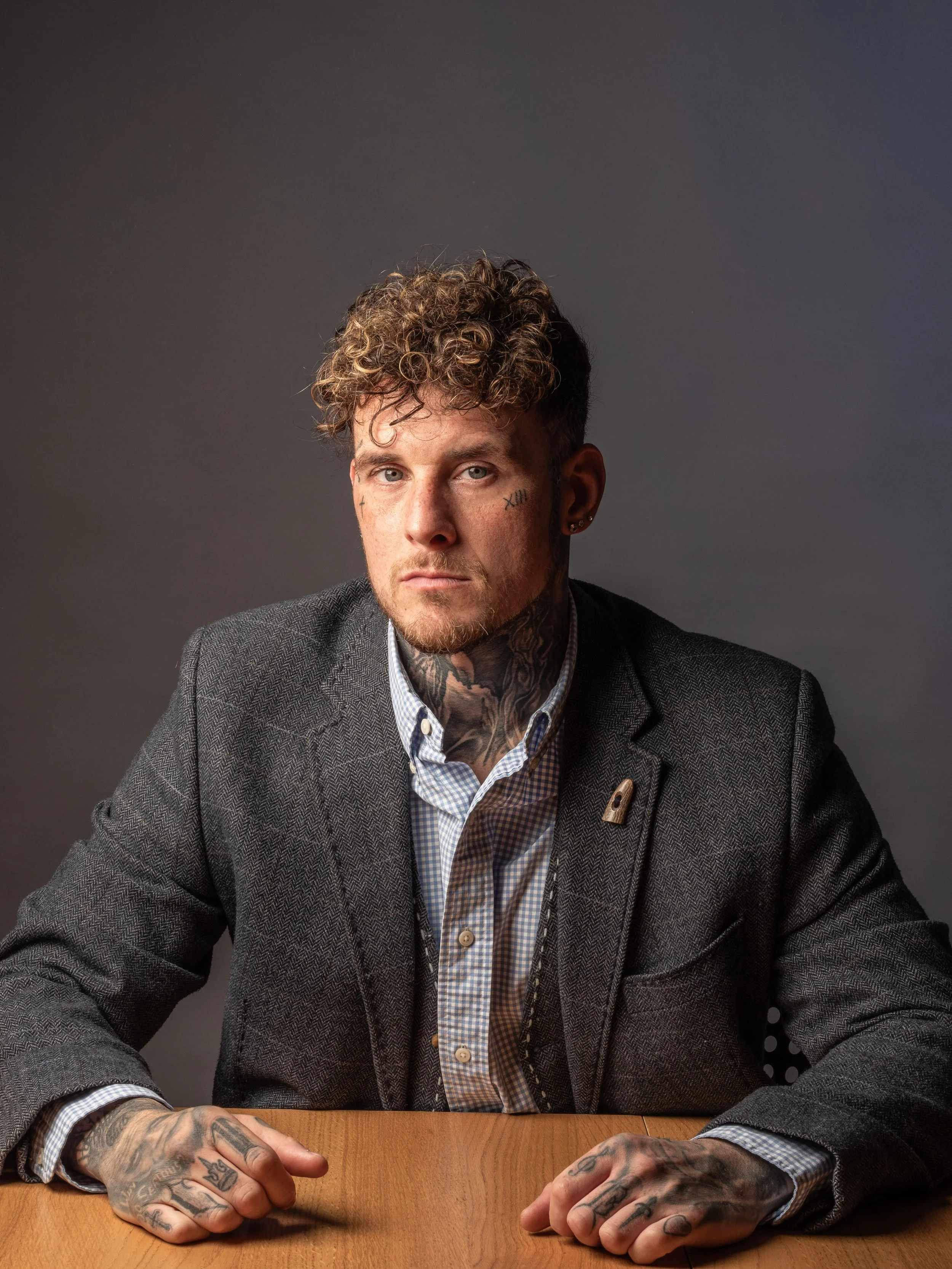 A man with curly hair, tattoos on his neck and hands, wearing a gray blazer over a checkered shirt, sitting at a wooden table against a gray background.