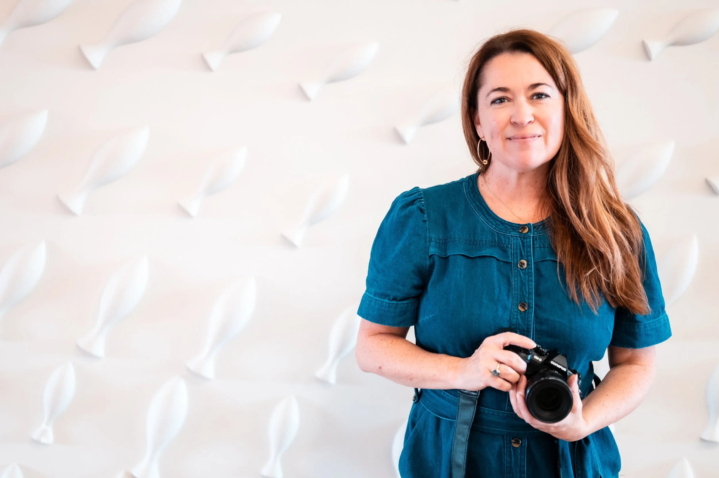 A woman with shoulder-length brown hair wearing a blue dress, holding a camera, standing in front of a white wall with 3D floral designs.