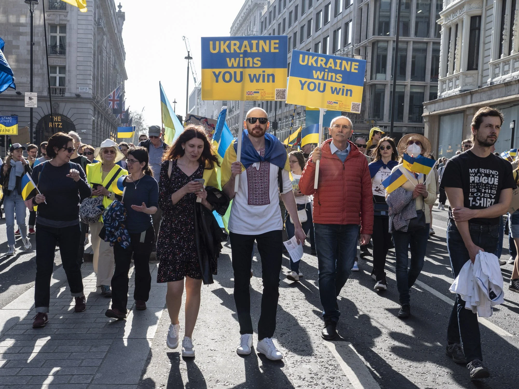 A crowd of people marching on a city street, holding Ukrainian flags and signs that say "UKRAINE wins YOU win." Some are wearing yellow vests and sunglasses, with tall buildings in the background.