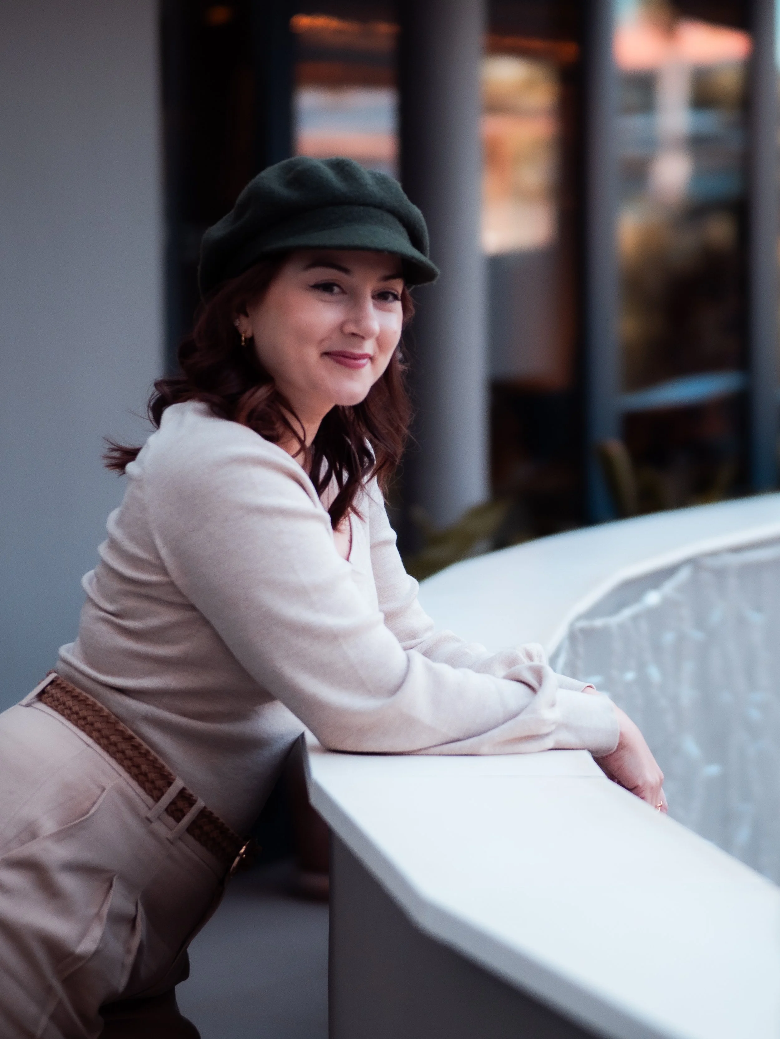 A woman with brown hair wearing a dark green beret, off-white sweater, and beige pants with a brown belt, leaning on a white curved railing indoors with large windows and warm lighting in the background.