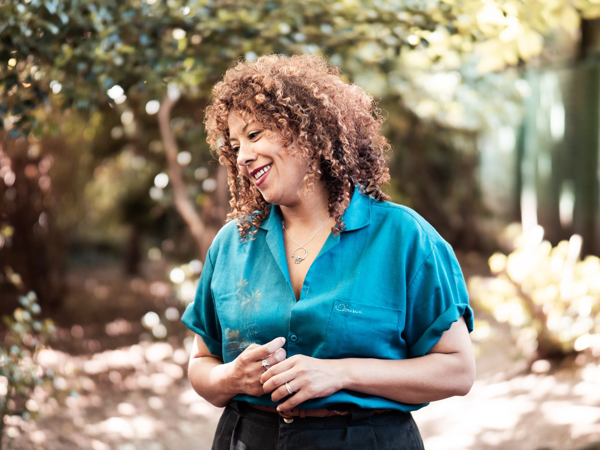 A woman with curly hair wearing a turquoise blouse, standing outdoors with trees and sunlight in the background, smiling and looking down.