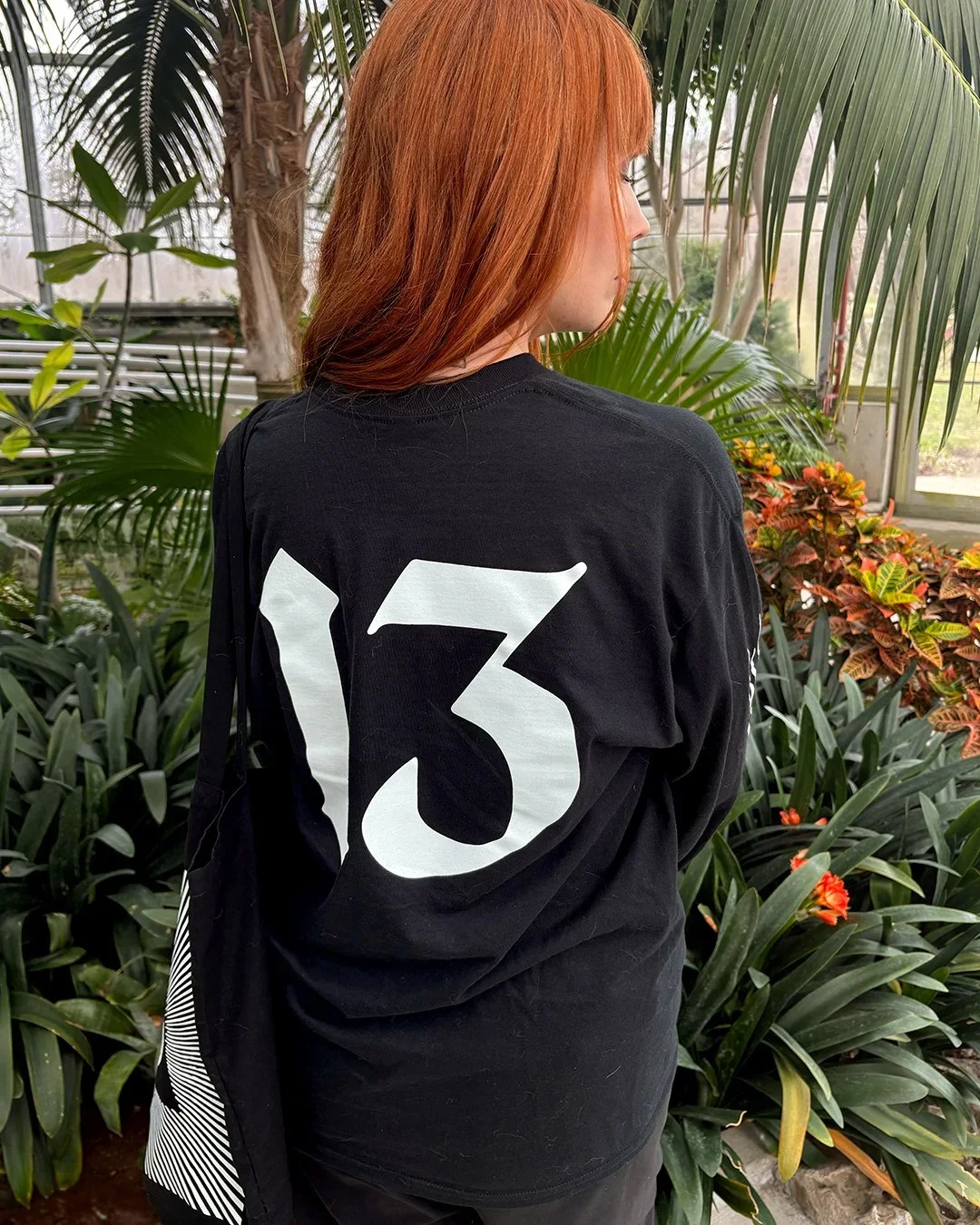 A woman with red hair wearing a black shirt with a large white number 13 on the back, standing in a greenhouse filled with green plants and sunlight.