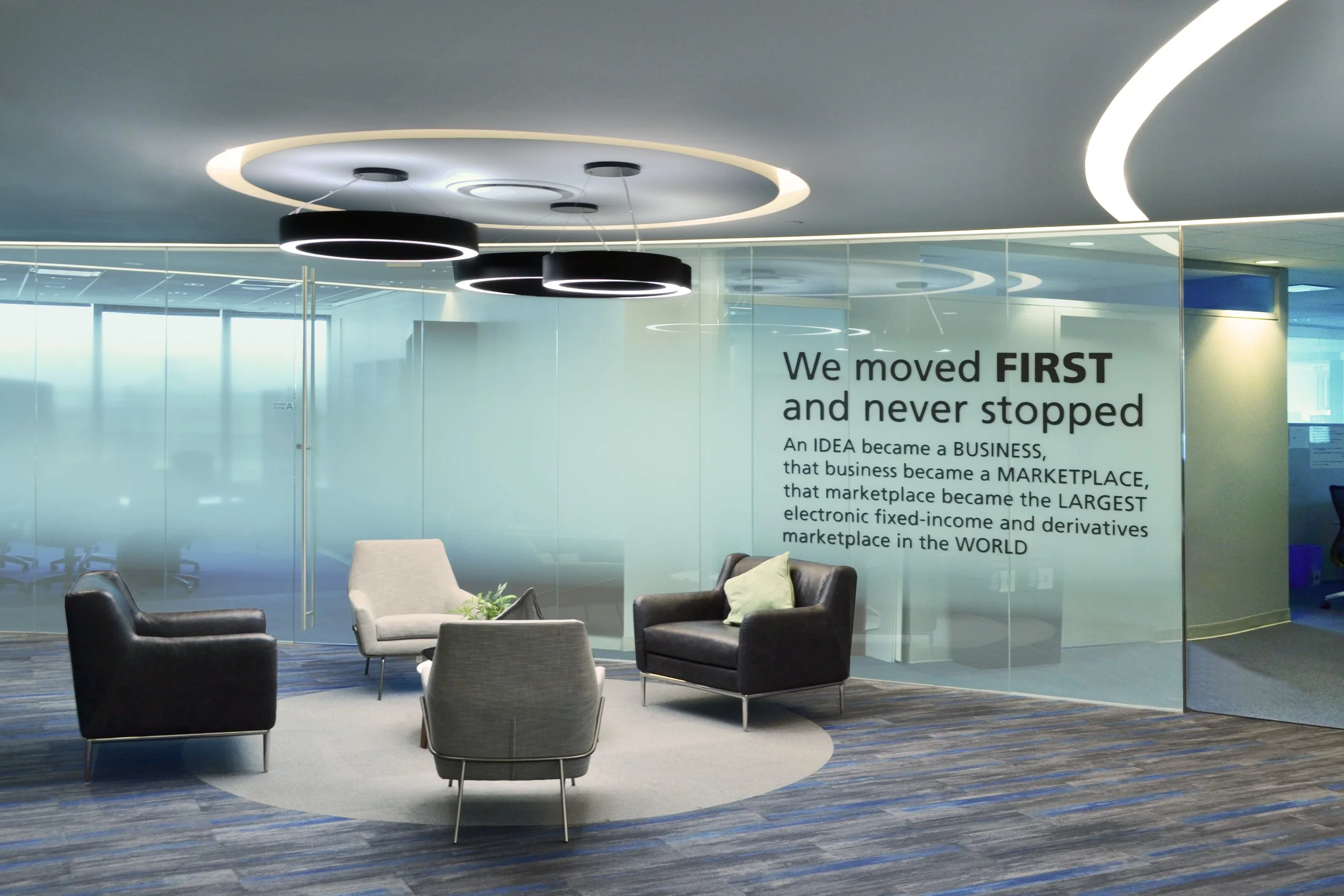 Modern office lounge area with chairs, glass wall with motivational quote, and circular ceiling lights.