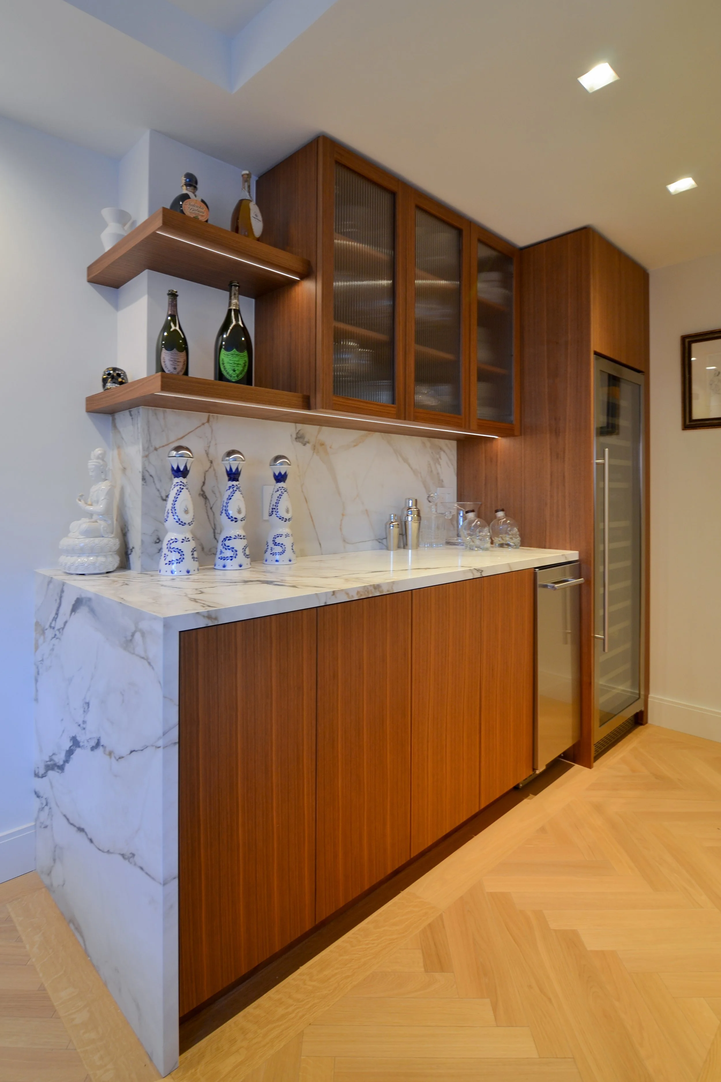 Modern home bar with wooden cabinets, marble countertop, glass shelves, and decorative bottles.