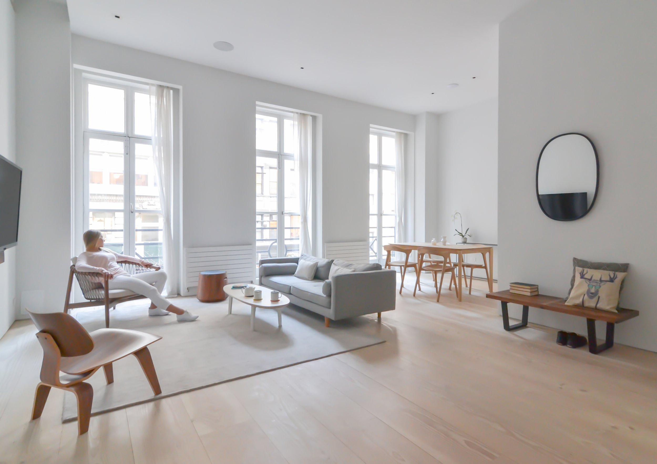 Modern living room with minimalistic design, featuring large windows, a gray sofa, wooden chairs, a coffee table, and a dining area with a table and chairs. A person is seated near the window.