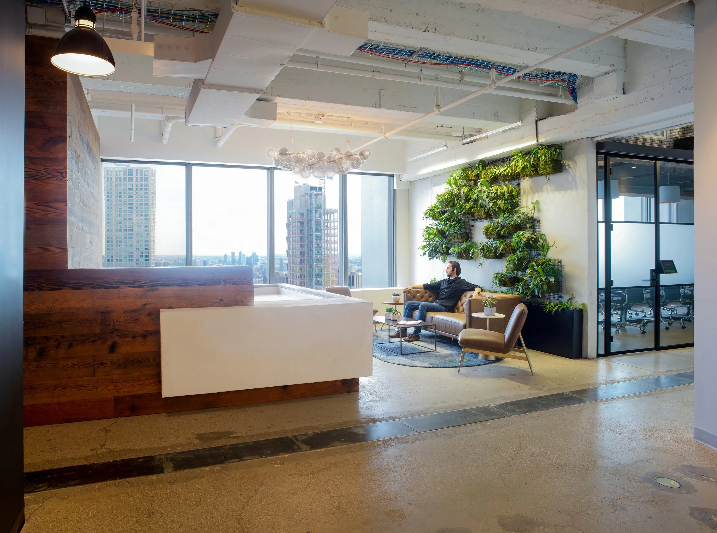 Modern office lounge area with large windows, green vertical plant wall, sofa and chairs, wooden desk, and city view.