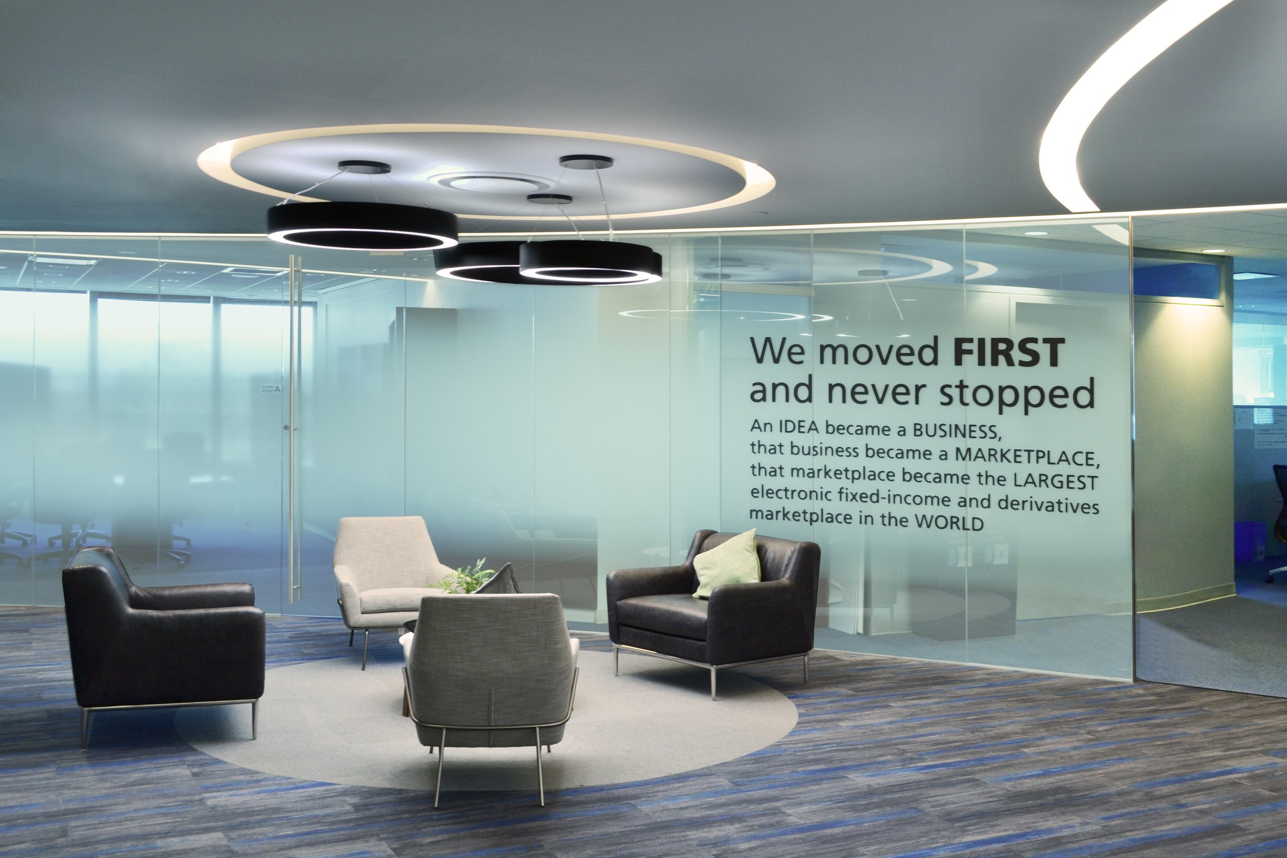 Modern office lobby with gray and black armchairs, circular ceiling lights, and motivational quote on a glass partition.