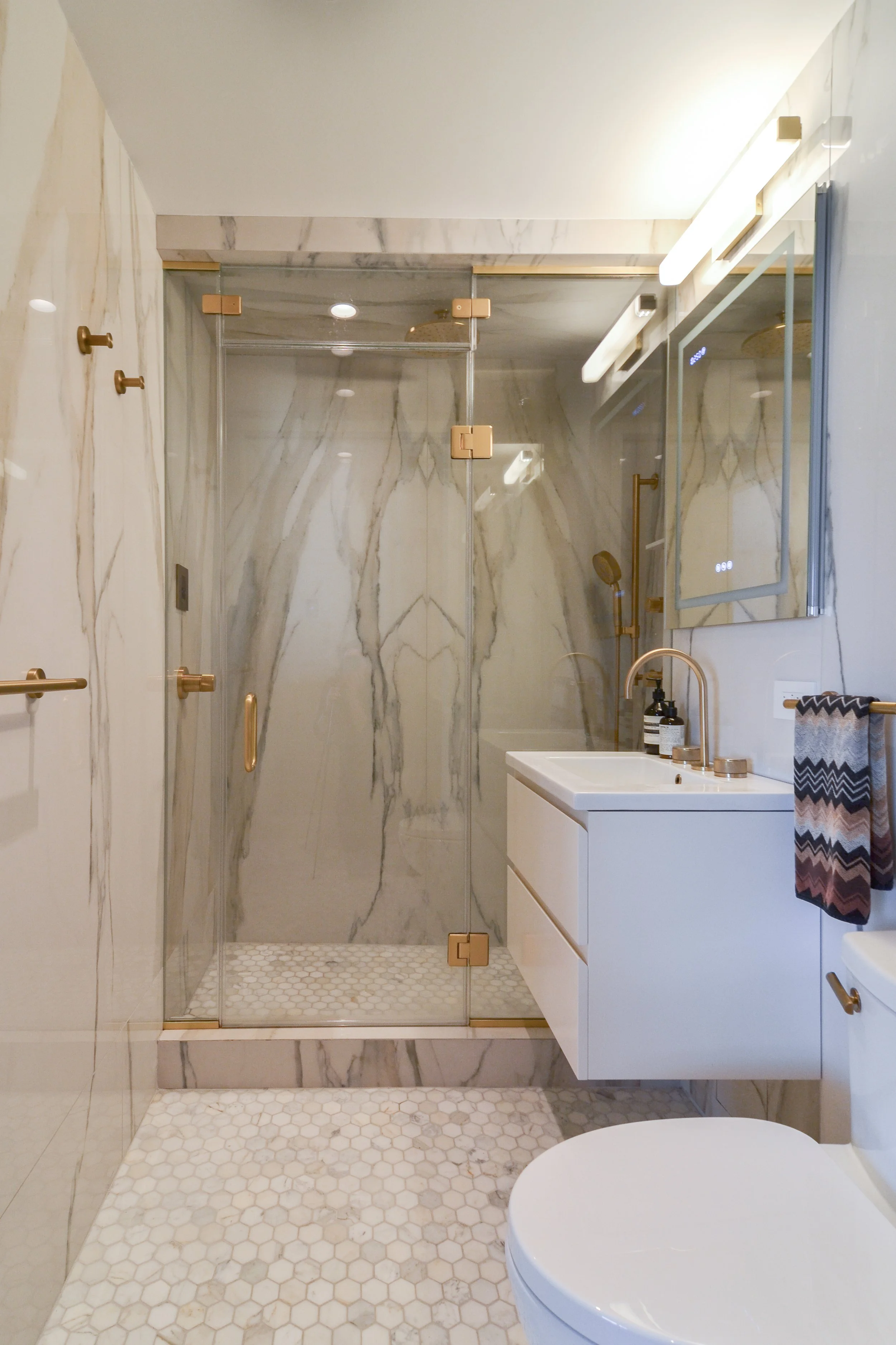 Modern bathroom with glass shower, marble walls, gold fixtures, white vanity, and hexagonal floor tiles.