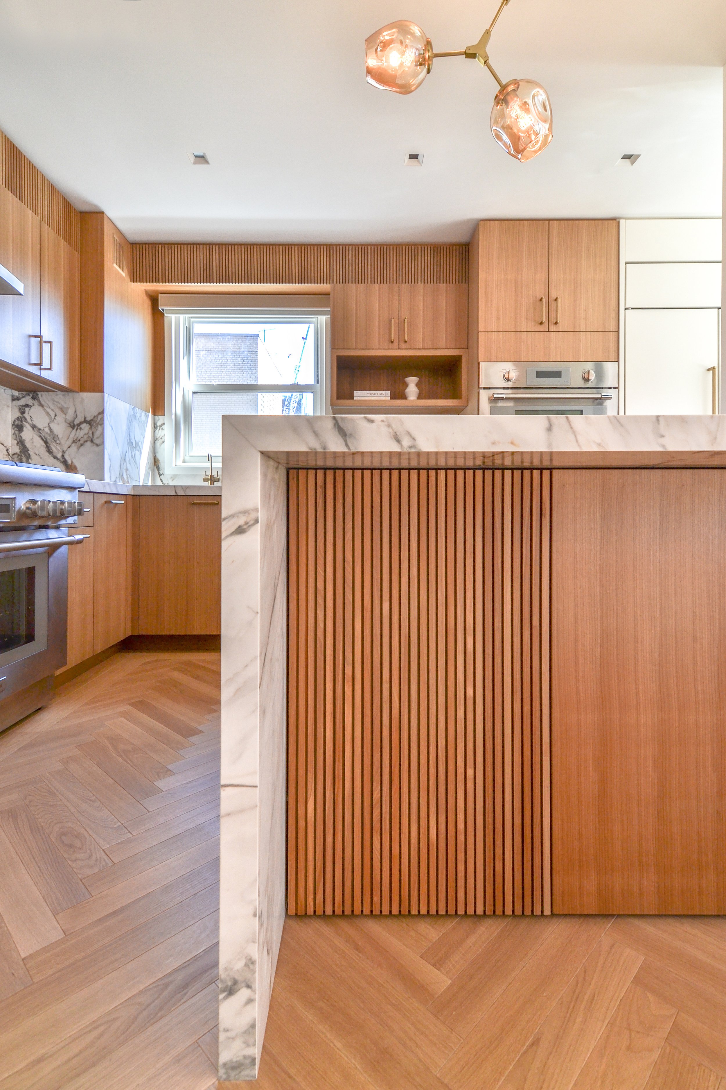 Modern kitchen with marble island, wooden cabinets, and herringbone wood flooring.