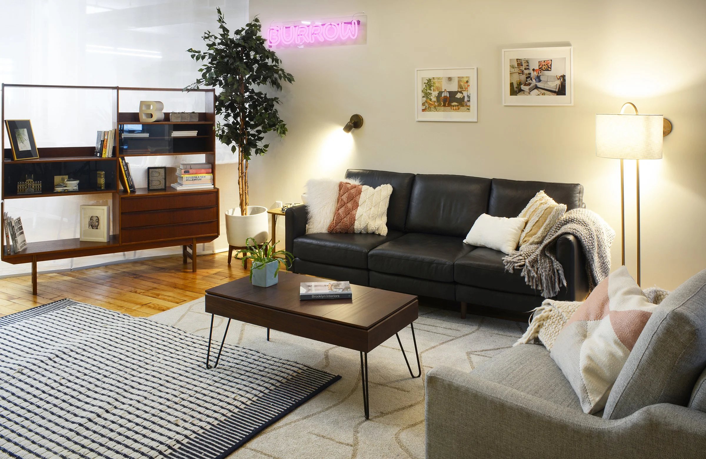 Stylish living room with black leather sofa, wooden coffee table, bookshelf, and neon sign. Features decorative pillows, plants, framed pictures, and floor lamp.