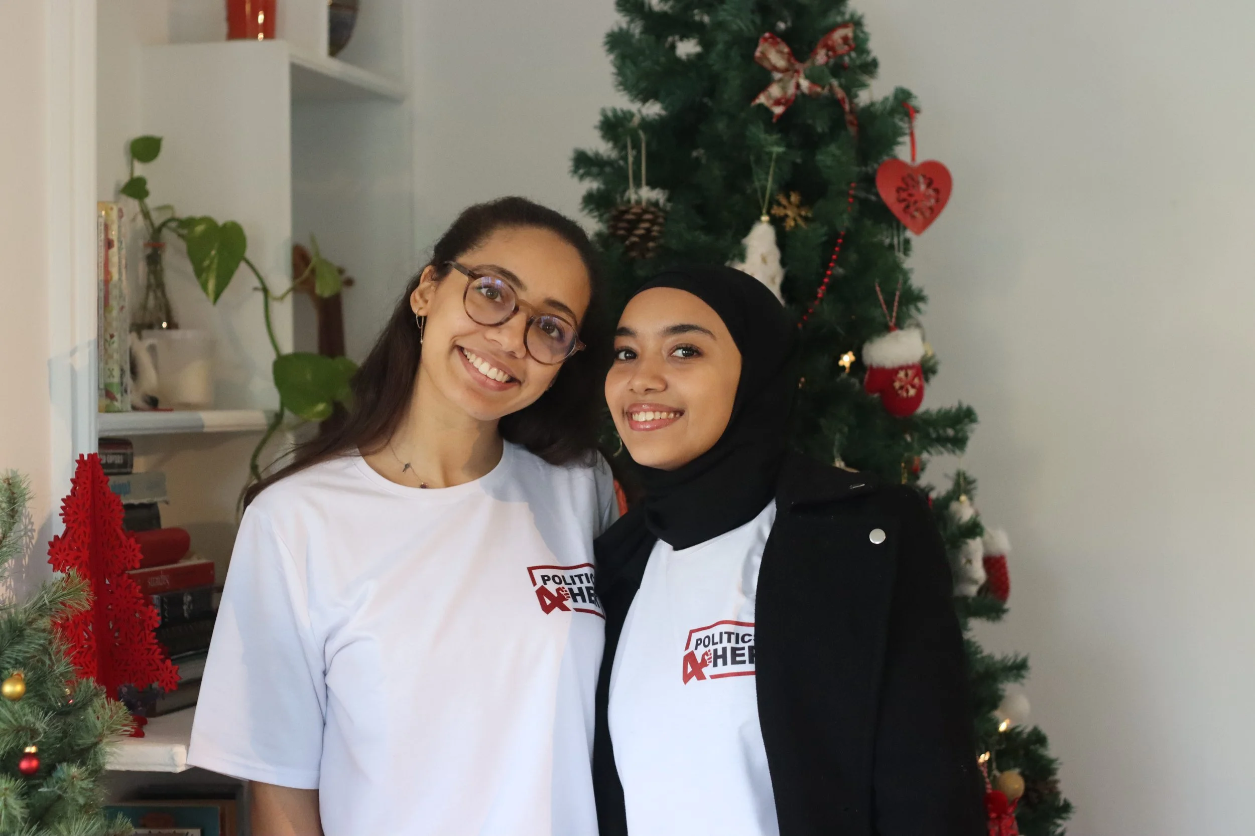Two young women standing close together near a decorated Christmas tree indoors. The woman on the left has long dark hair, glasses, and is wearing a white shirt with a Political 4Her logo. The woman on the right has a hijab, a black jacket, and a whi