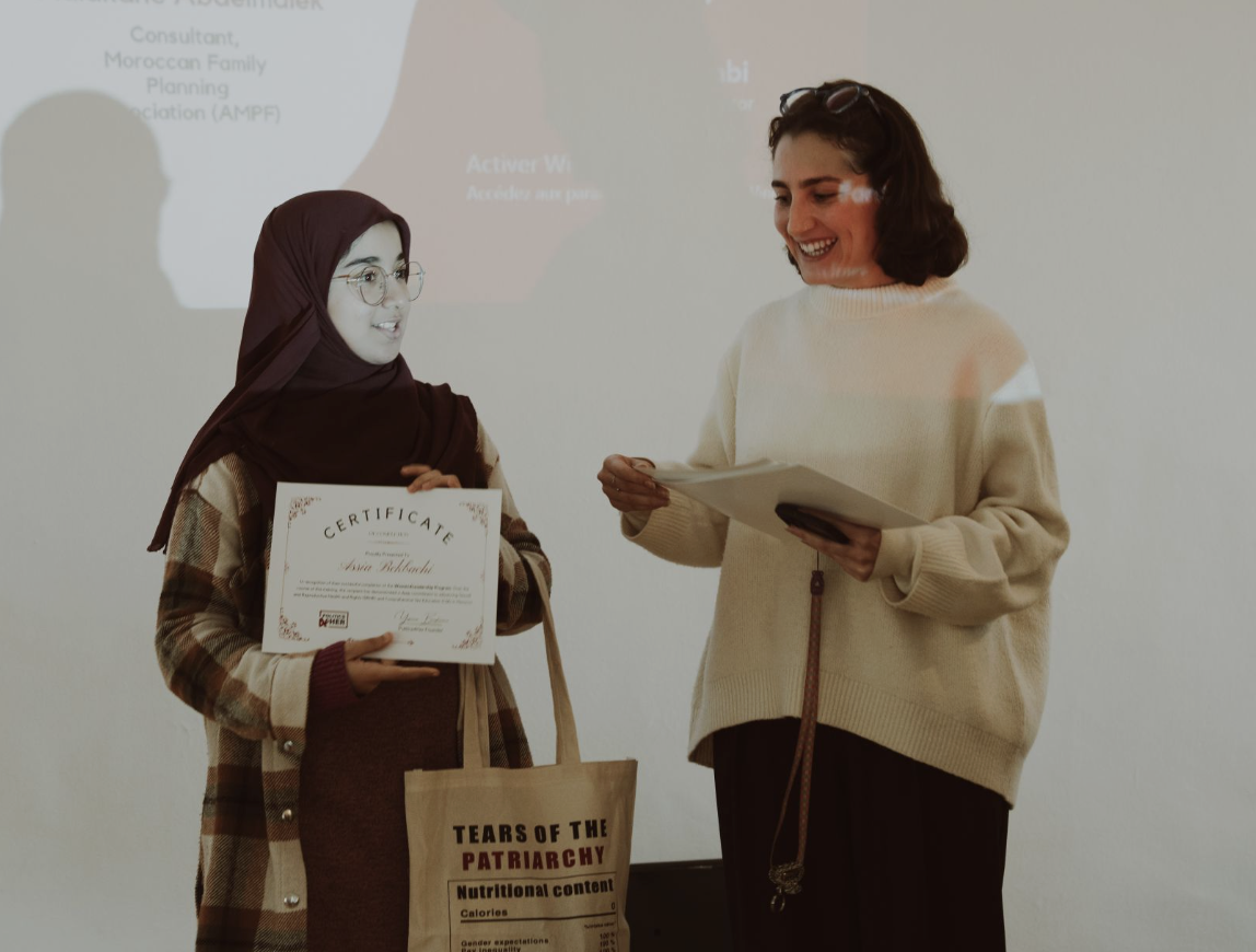 A woman in a beige sweater is presenting a certificate to another woman in a hijab, glasses, and a plaid coat, during an award ceremony.