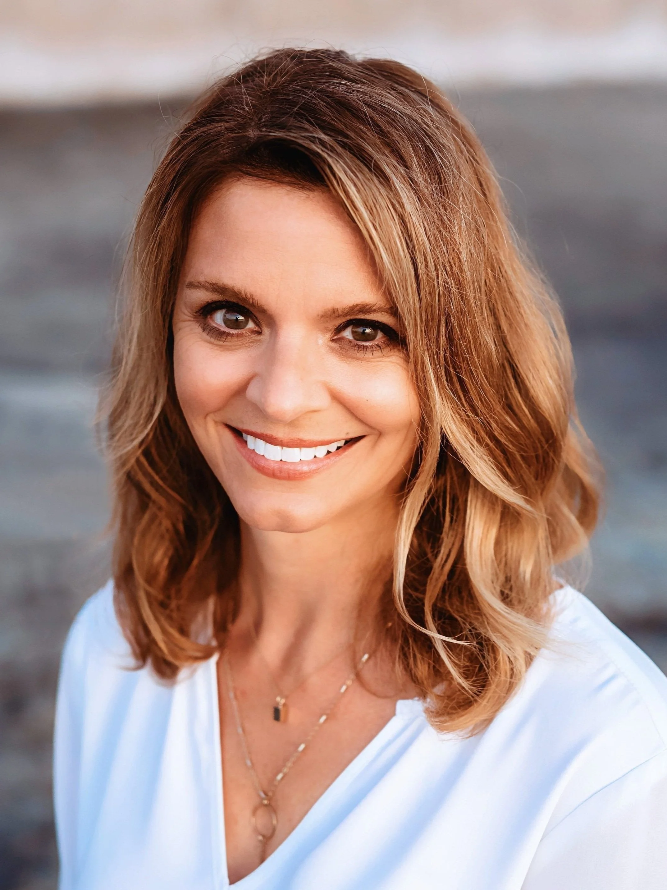 A professional woman with shoulder-length, light brown hair smiling at the camera, wearing a white top and layered necklaces, with a blurred background of water.