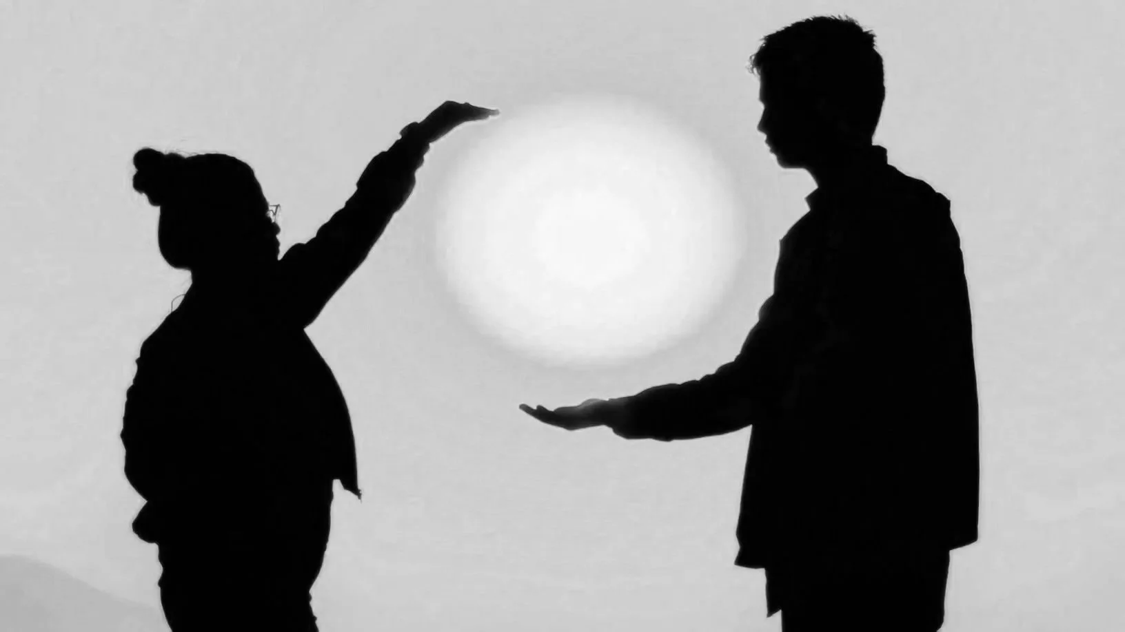Silhouettes of a woman and man against a bright circular background, with the woman holding an open hand above the man's hand, creating a visual of holding a glowing orb between them creating a common space for connection to occur.