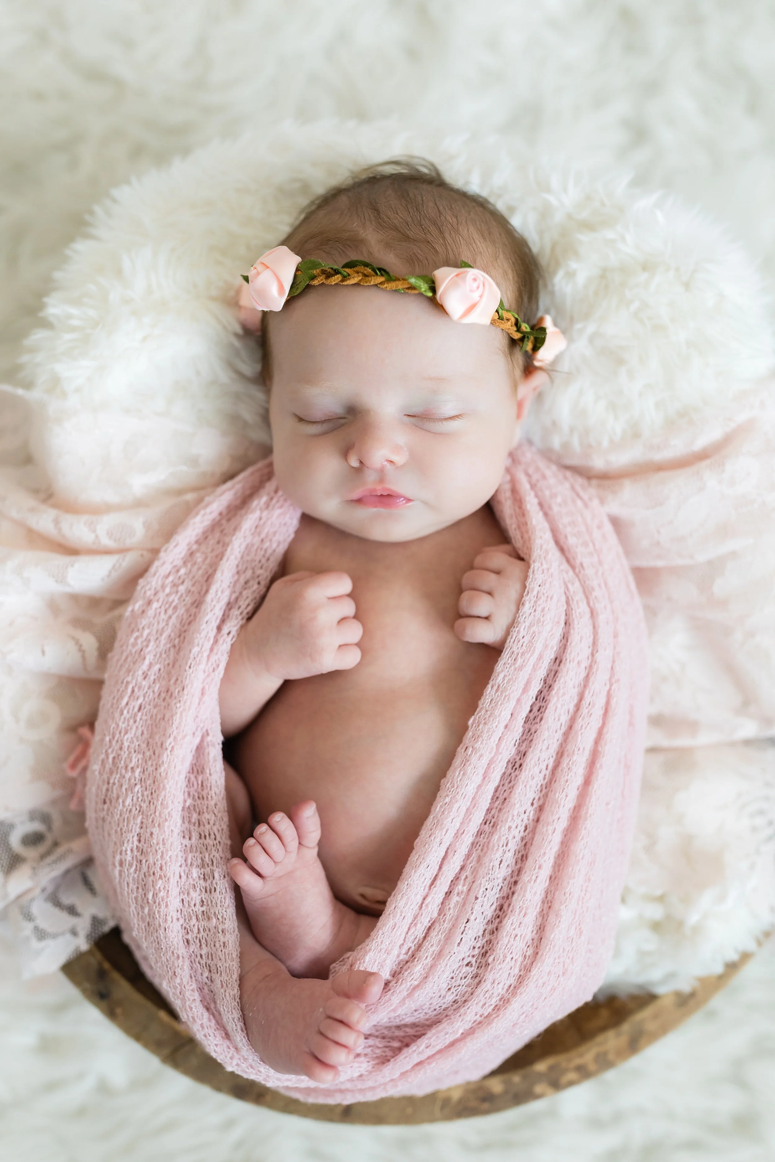 A sleeping baby wrapped in a pink knitted blanket, with a floral headband, lying on a fluffy white surface inside a round wooden container.