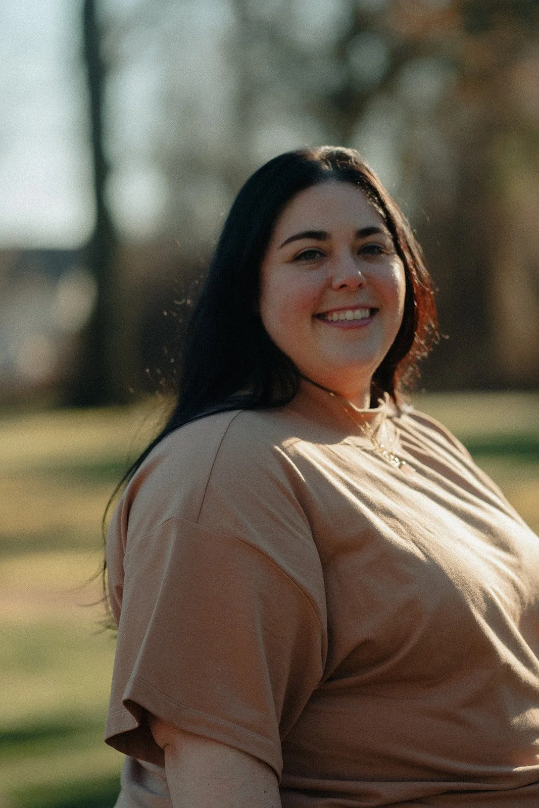 A young woman with long dark hair smiling outdoors on a sunny day, wearing a beige t-shirt and a choker necklace.