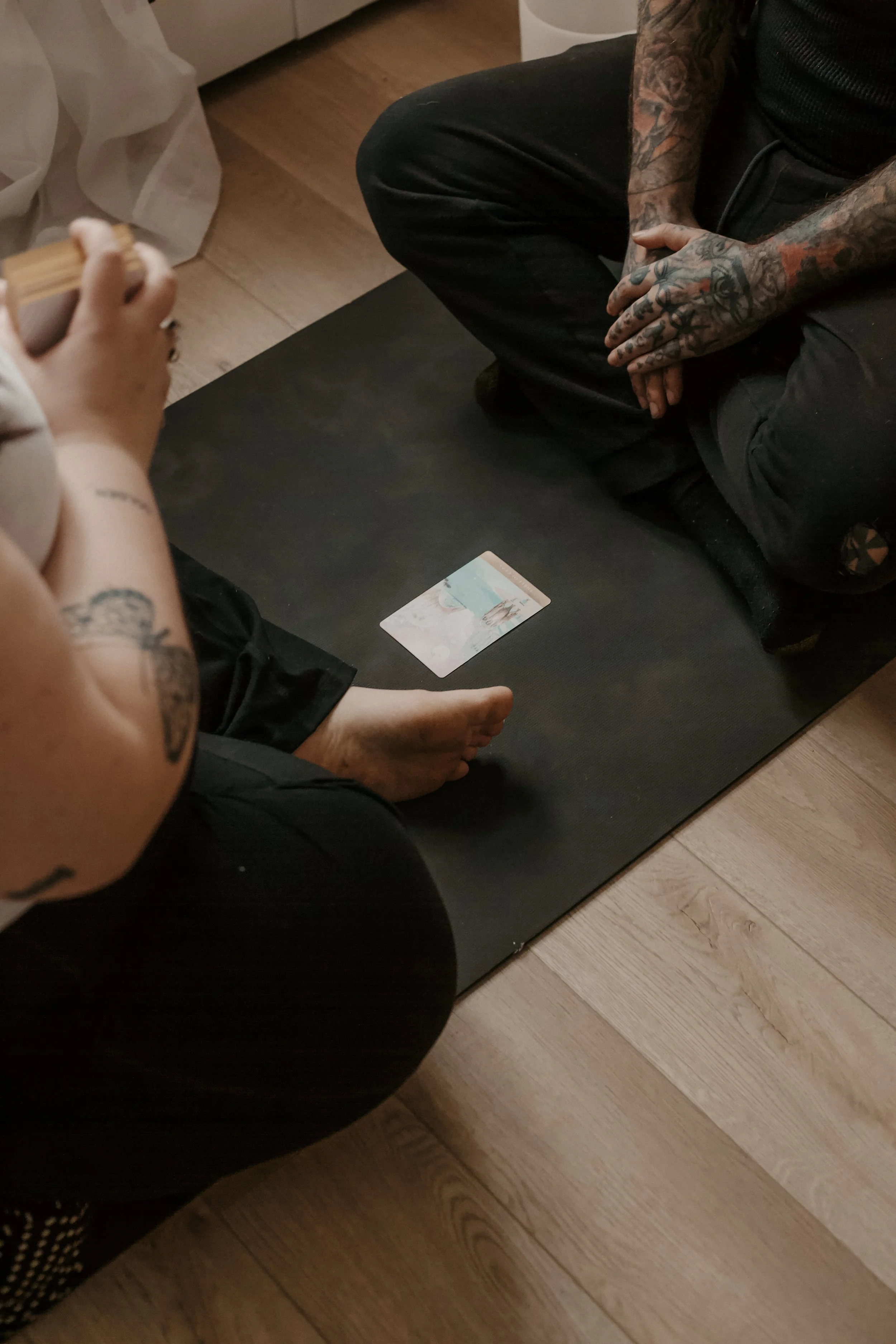 Two people sitting on the floor with a tarot card placed on a black mat between them. One person is barefoot and holding a deck of tarot cards, while the other person has tattoos on their arms and is sitting cross-legged.