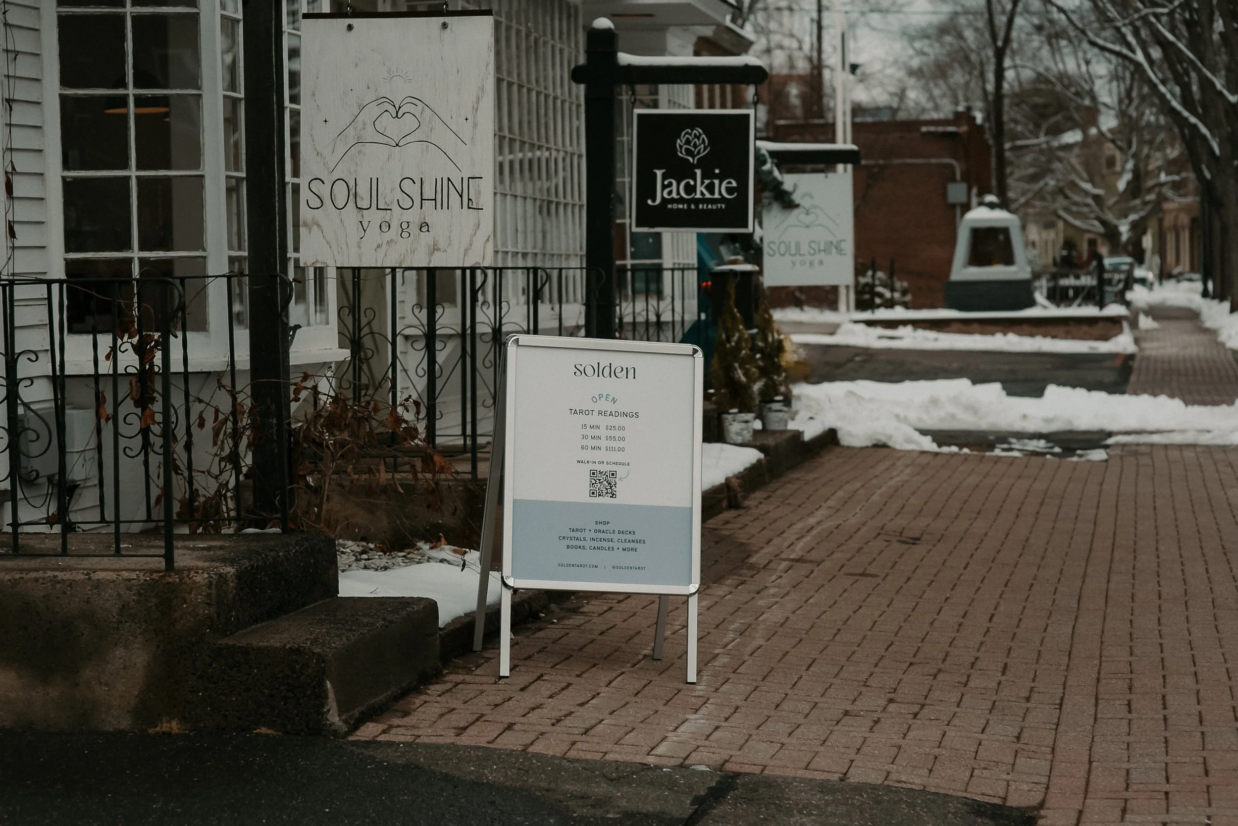 Sidewalk cafe with signs for Soulshine Yoga, Jackie Home & Beauty, and Solden offering tarot readings; snow on the ground with bare trees and buildings in the background.
