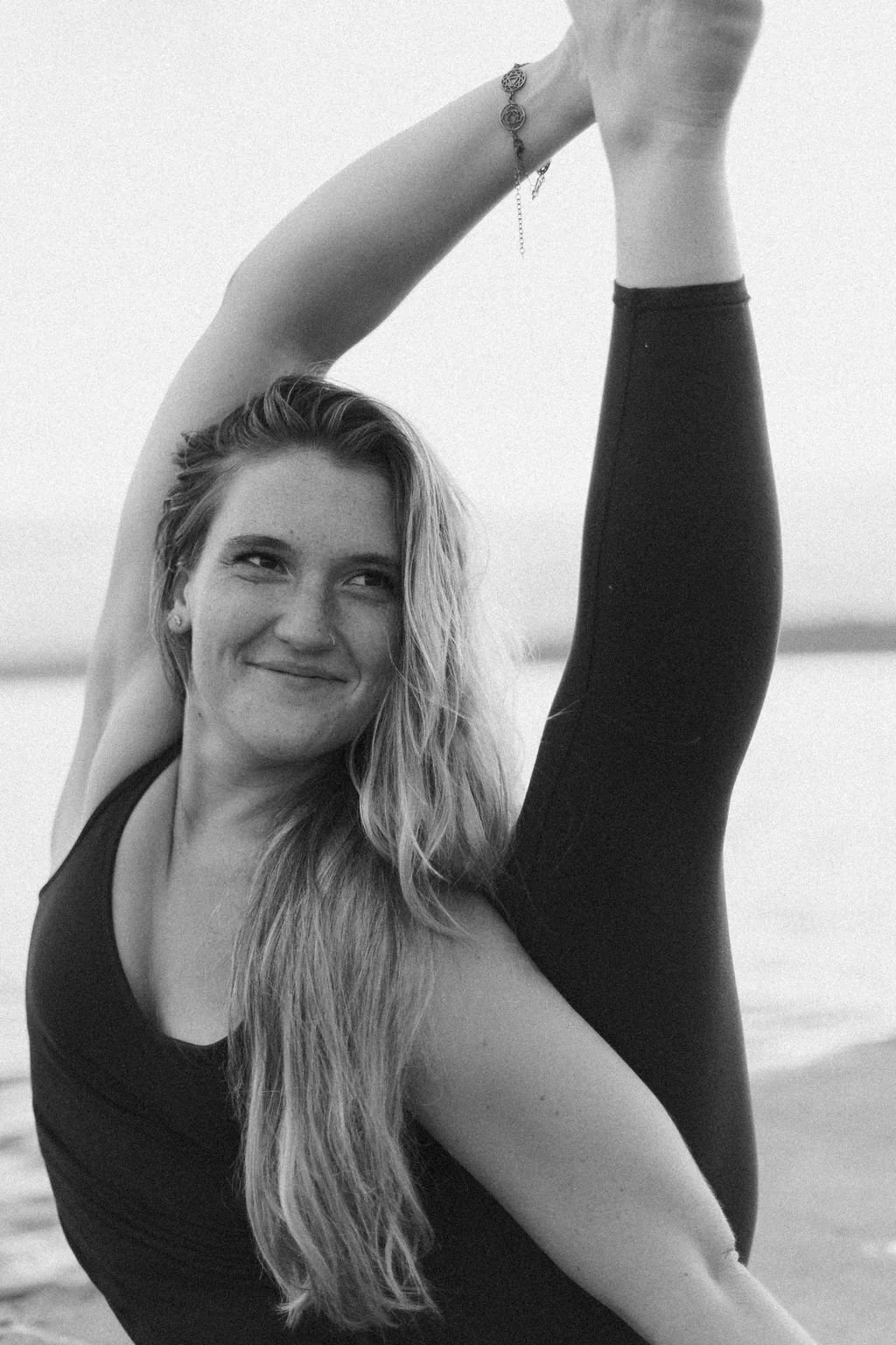 Woman practicing yoga on the beach, stretching her leg overhead with a playful expression on her face.