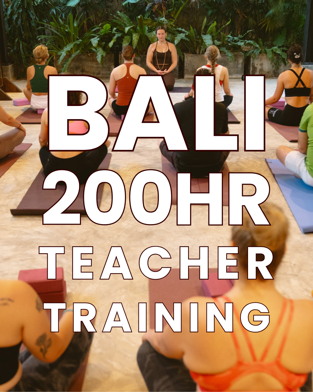 A group of people participating in a yoga or meditation class indoors with lush green plants in the background. A teacher is leading the session while seated on a mat at the front, with students sitting cross-legged on mats facing her.