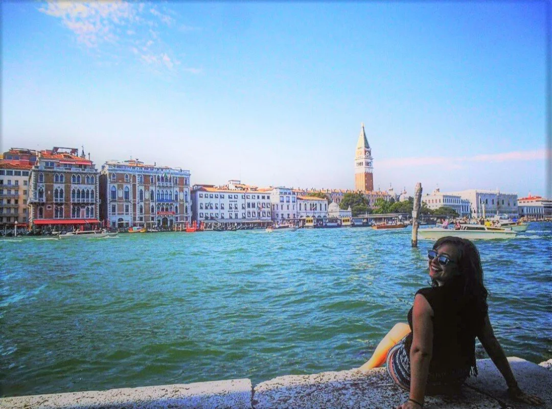 A woman sitting by a water canal in Venice, Italy, smiling and wearing sunglasses, with historic colorful buildings and the clock tower in St. Mark's Square in the background.