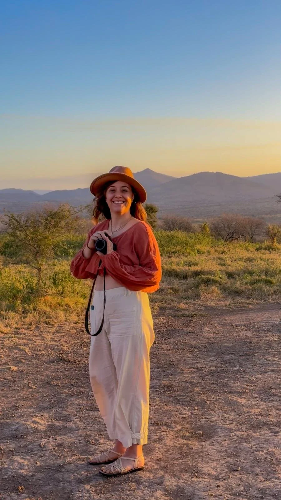 A woman standing outdoors on a dirt path, smiling and holding a camera, with mountains and a sunset sky in the background.