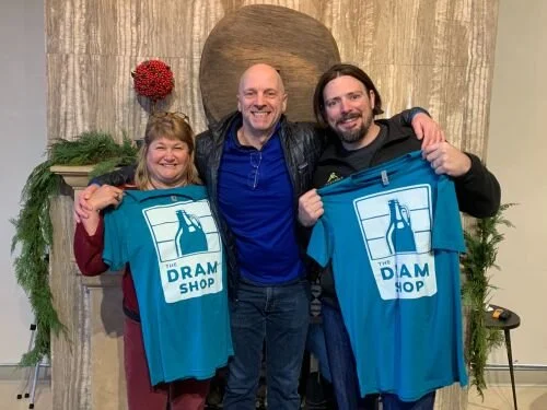 Three people smiling and holding blue shirts with a beer bottle and the text 'The Dram Shop' in front of a fireplace mantel decorated with red flowers and greenery.
