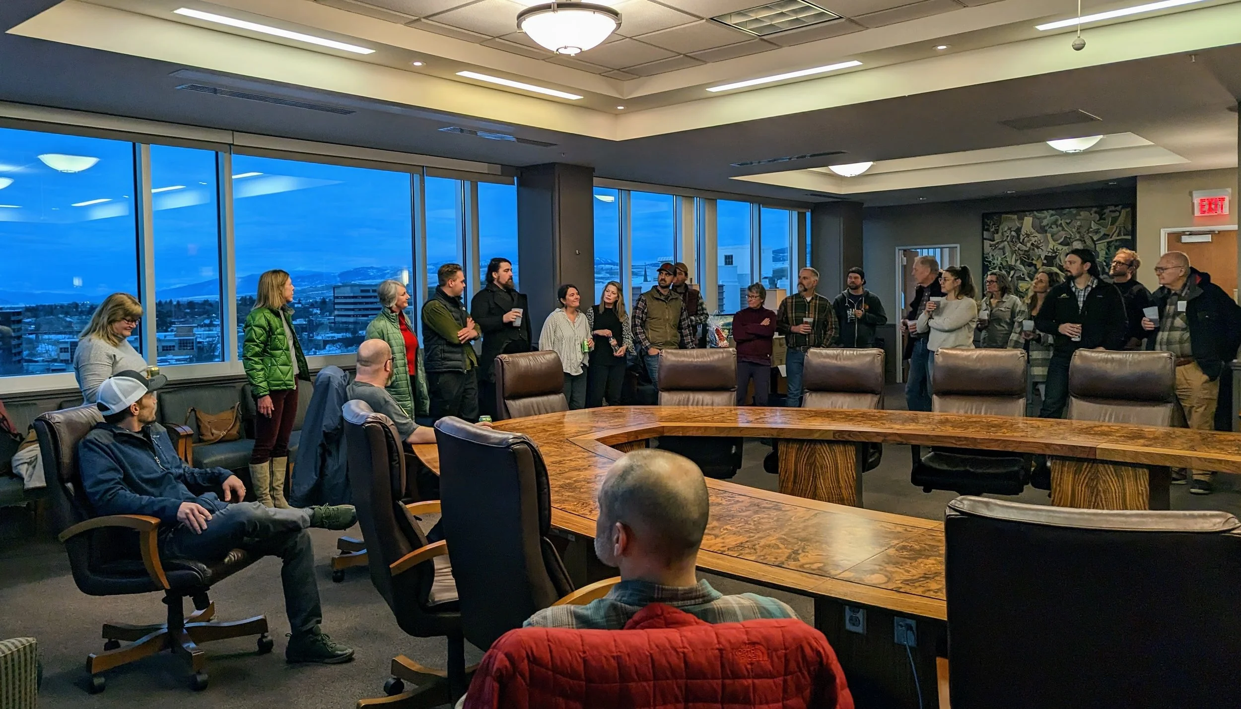 Group of people gathered in a conference room with large windows, some standing along the windows and some seated, during dusk. A wooden conference table is in the foreground.