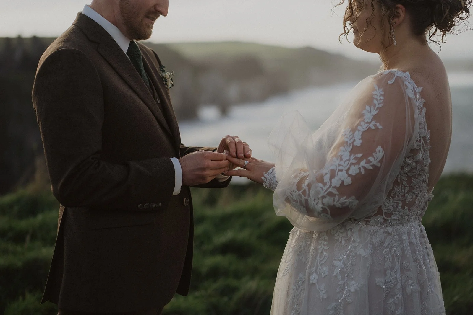 A groom puts his brides wedding ring on in front of Dunluce Castle in Northern Ireland. There are cliffs and the sea in the background.