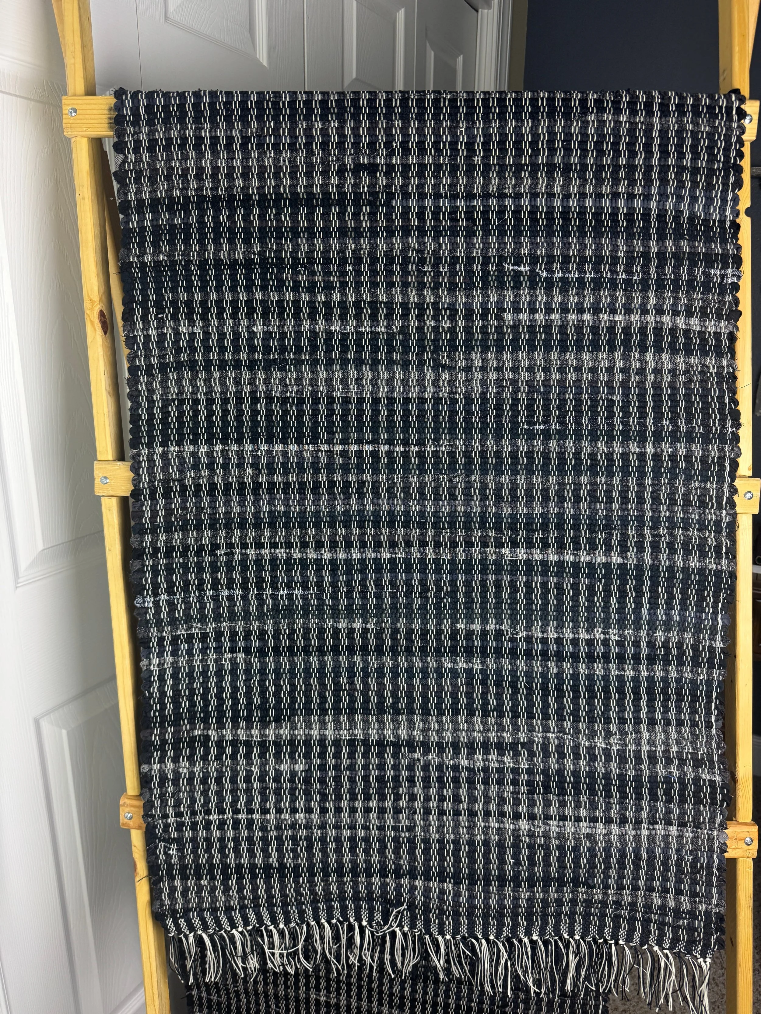 Black handwoven rag rug hanging on rack showing weave and fringe