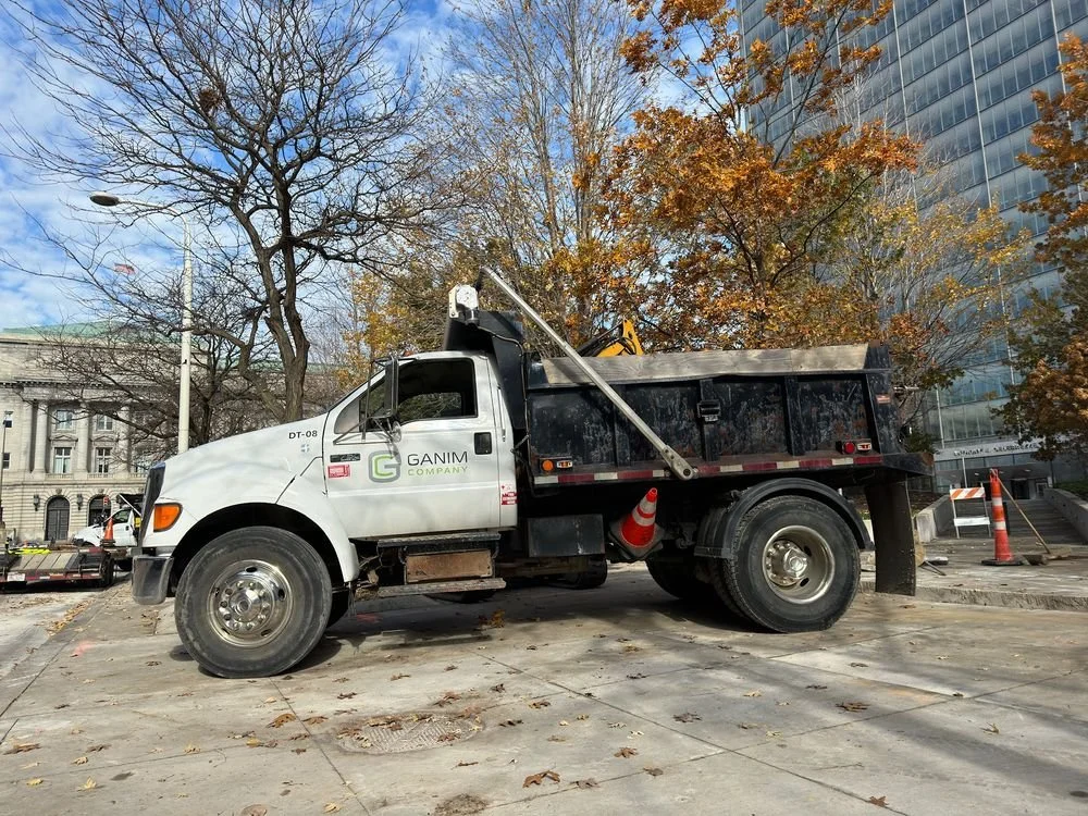 A white and black construction truck parked on a city street with autumn trees and buildings in the background.
