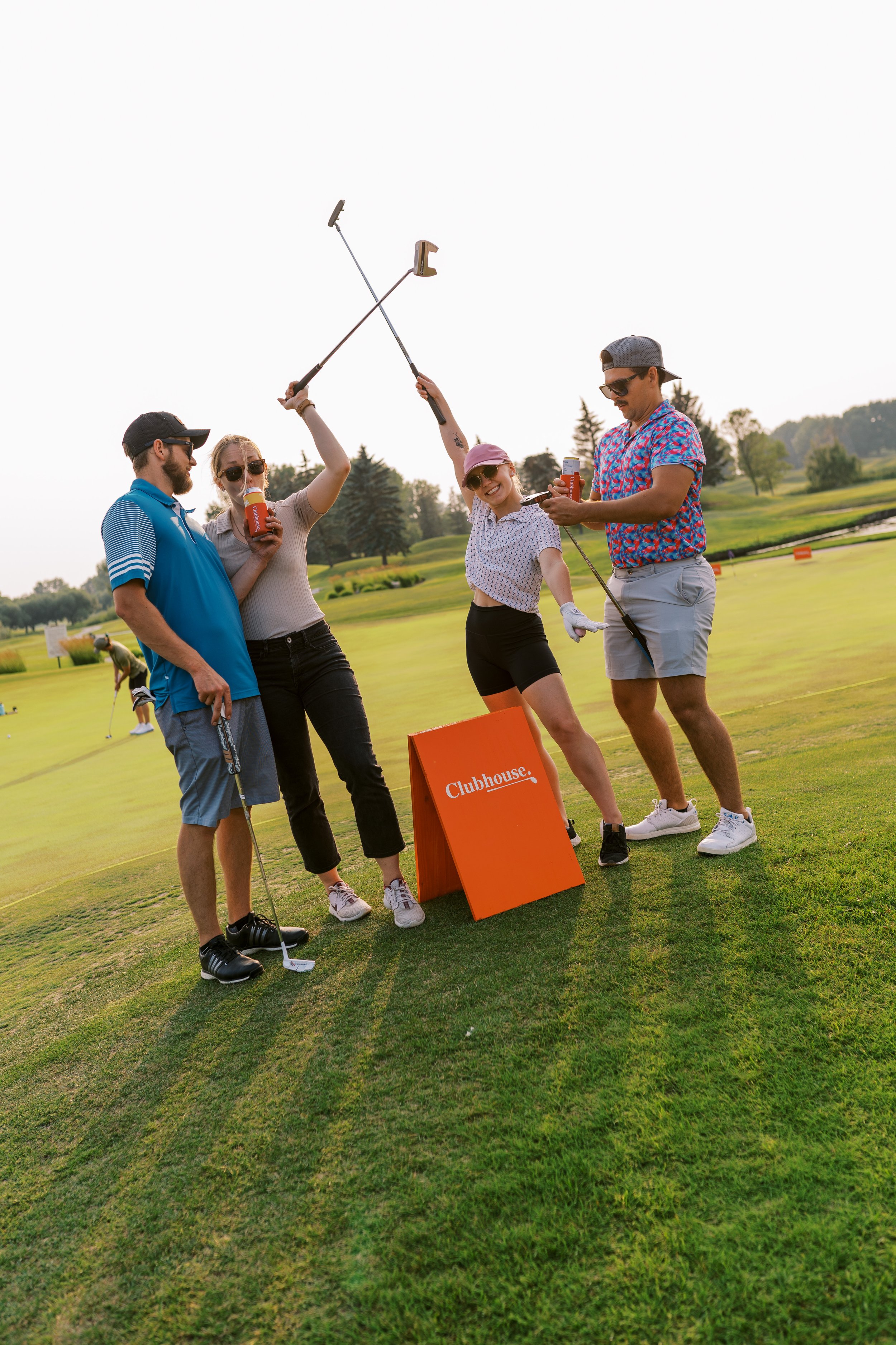 Group of friends at a golf course celebrating with drinks, holding golf clubs, with a club house sign in the foreground and trees in the background.