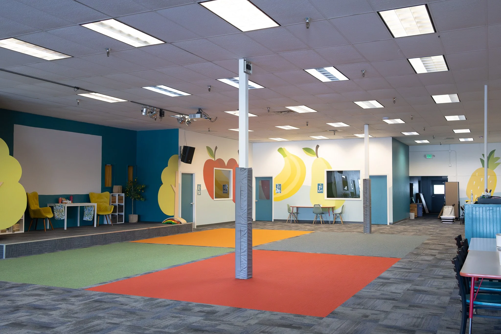 Colorful indoor play area with a stage, chairs, and murals of fruits on the walls.