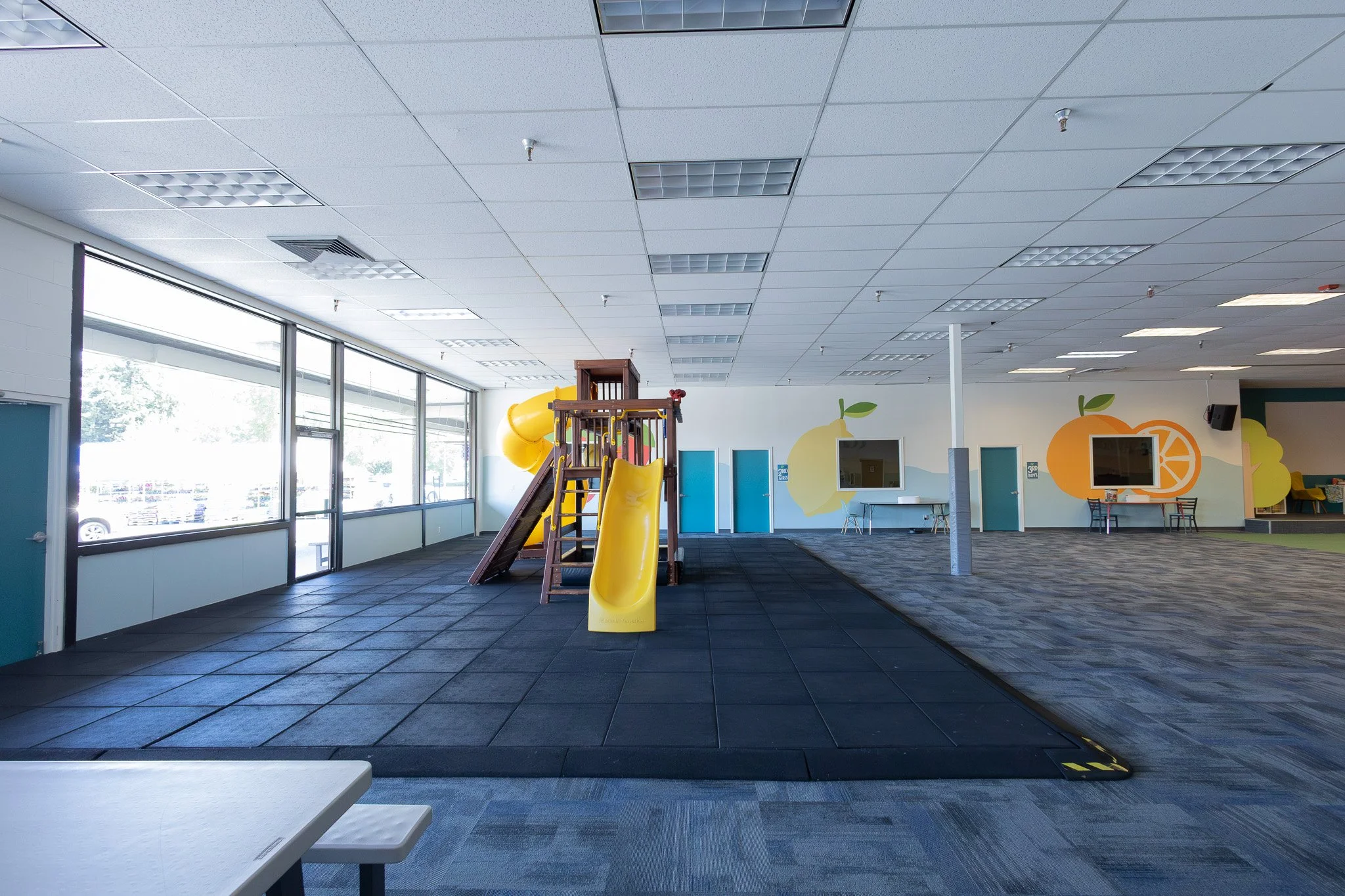 Indoor children's play area with a yellow slide, a wooden climbing structure, and colorful wall decorations of fruits and trees.