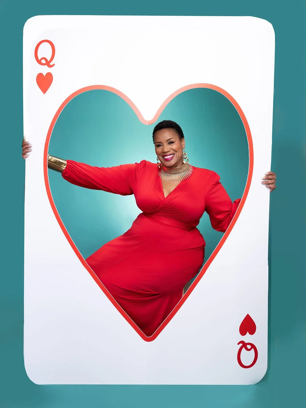 A woman in a red dress posing through a large Queen of Hearts playing card cutout.