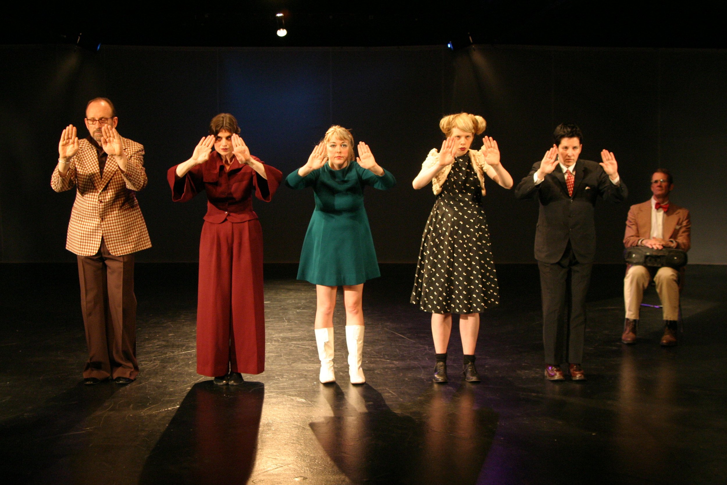 five dancers in a line facing front, hands flat out in front of them, a man sitting in a chair at the right with a boombox on his lap