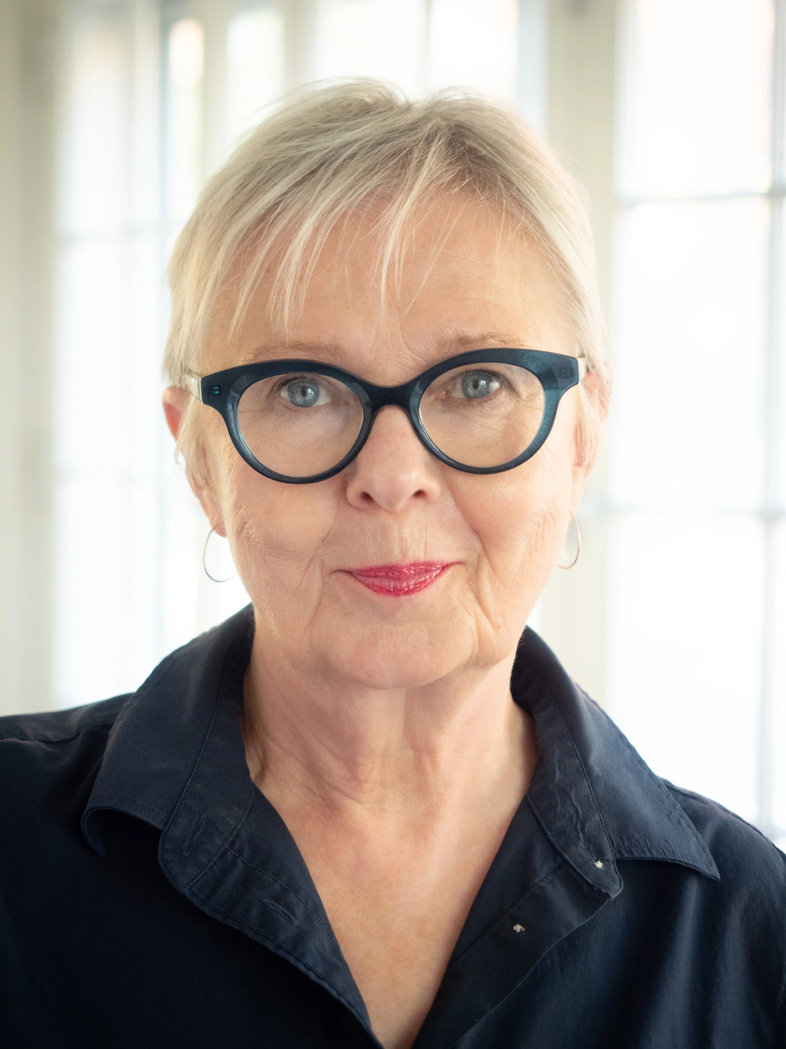 A head-and-neck portrait of a light-skiunned woman with short blonde hair and blue eyes. She wears a navy shirt and dark glasses and looks directly at the camera with a slight smile. The background is softly out of focus, w/a white window behind her.
