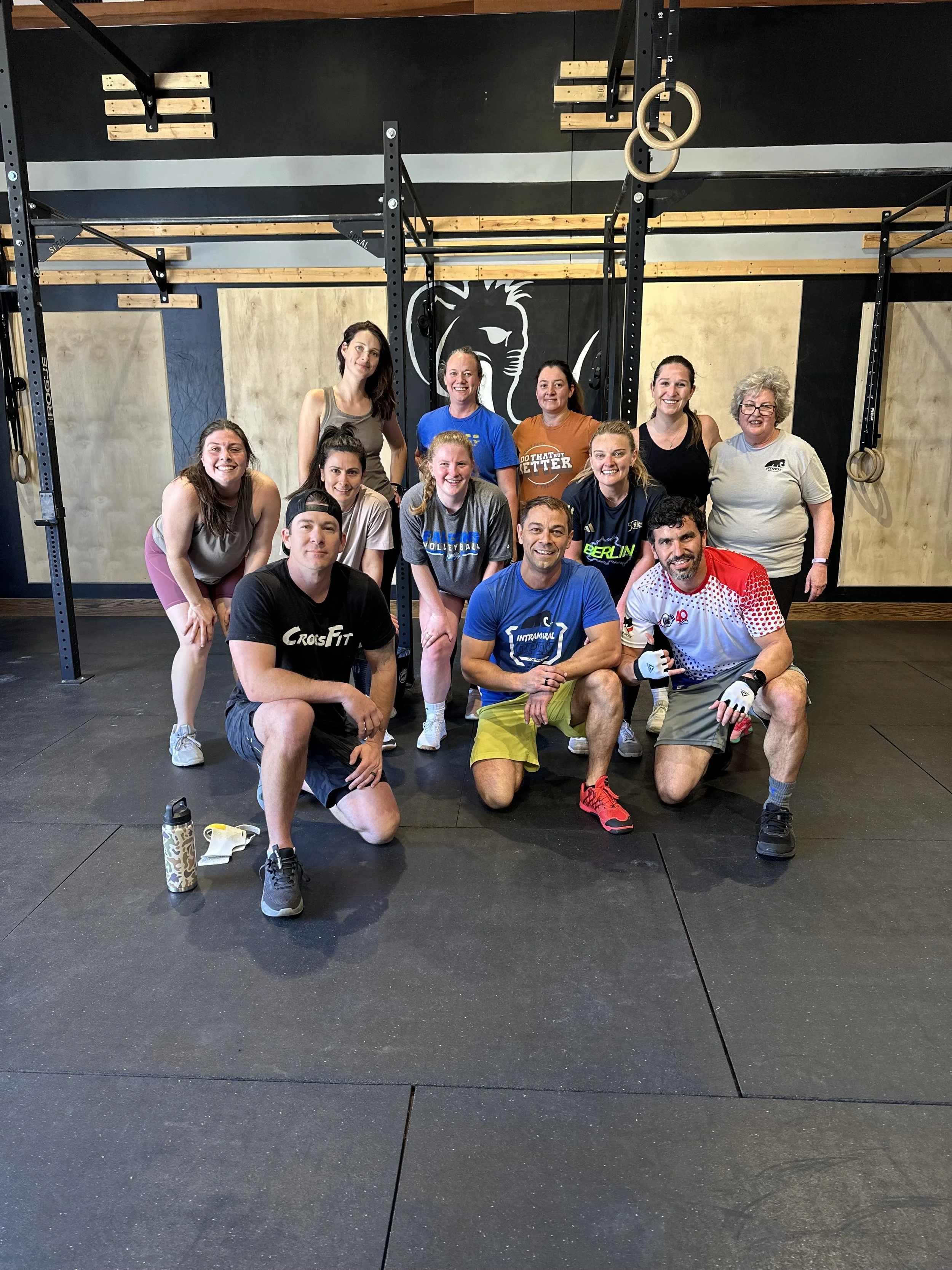 A group of eleven of our members at CrossFit Woolly Mammoth posing together, some kneeling and some standing, with gym equipment and a lion logo on the wall behind them.